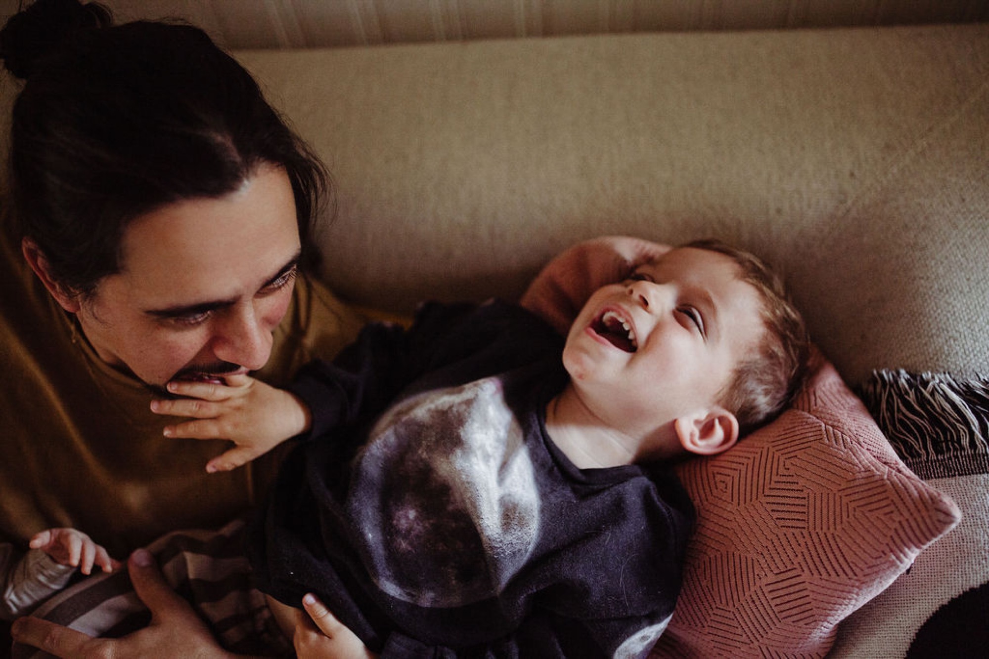 Mother gazes down at her laughing infant lying on a neutral-toned sofa, their joyful moment bathed in warm, soft natural light