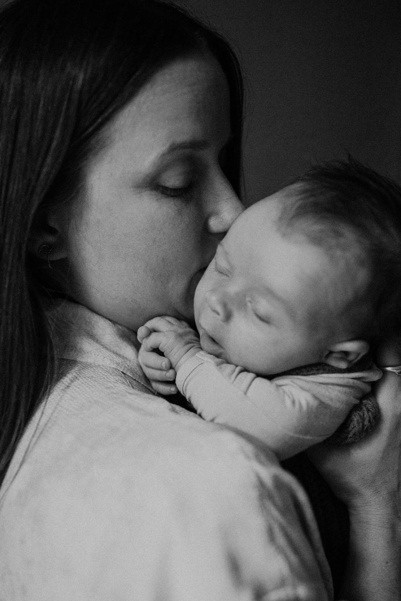 Mother kisses her sleeping newborn's forehead in tender profile, soft natural light sculpting their peaceful faces