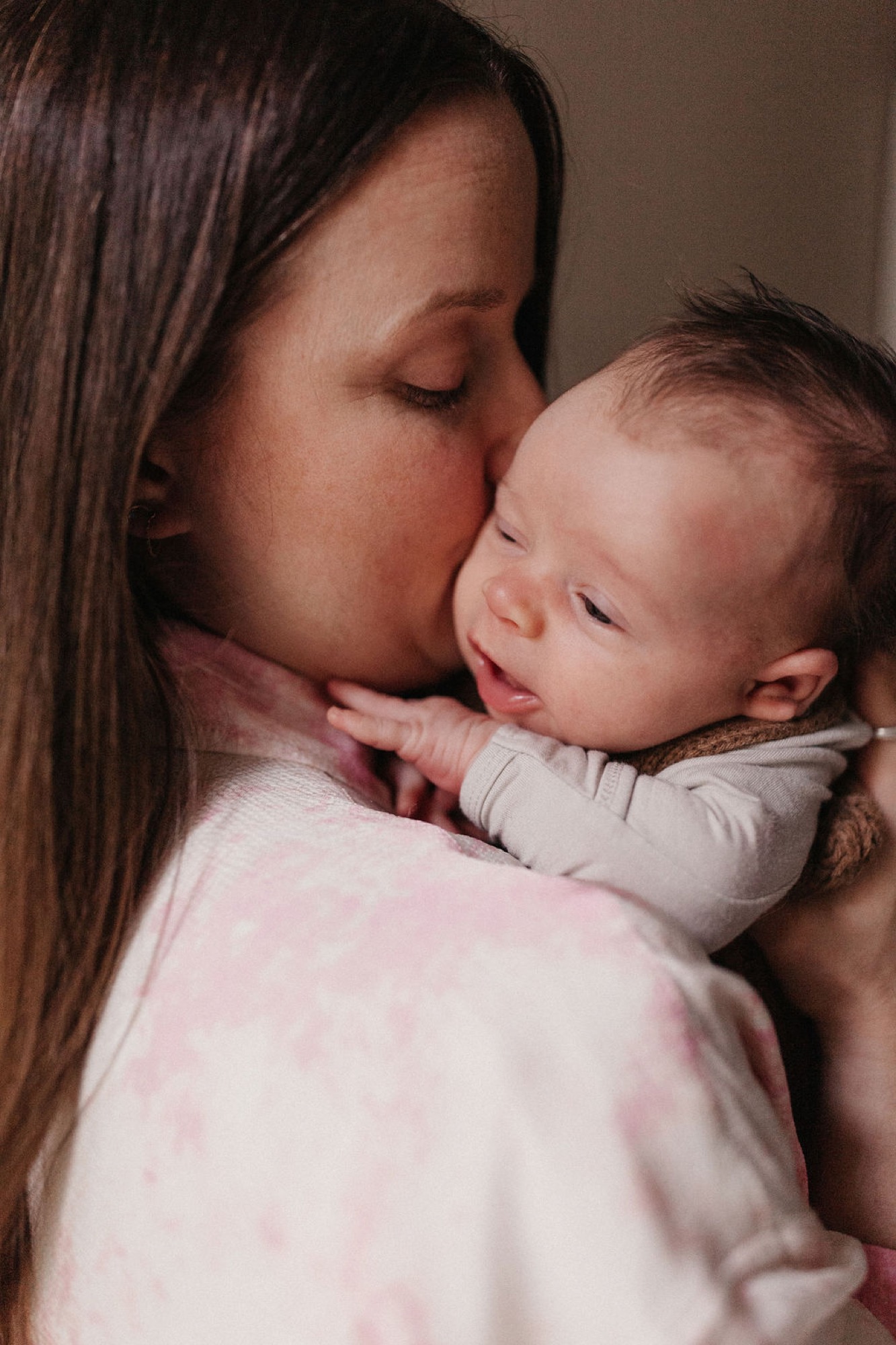 Mother in soft pink holds her newborn close, kissing the baby's forehead in warm, intimate indoor light