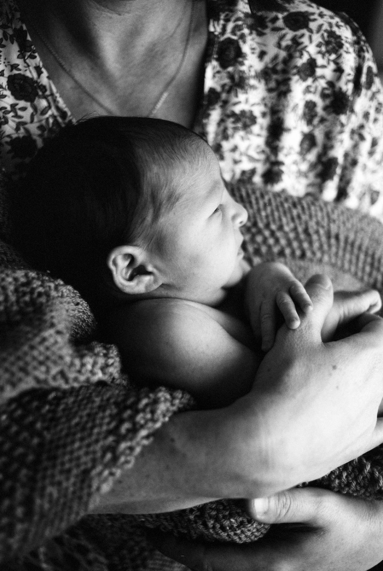 Newborn nestled against mother's chest, gazing upward with tiny hand reaching toward her textured sweater in intimate indoor light
