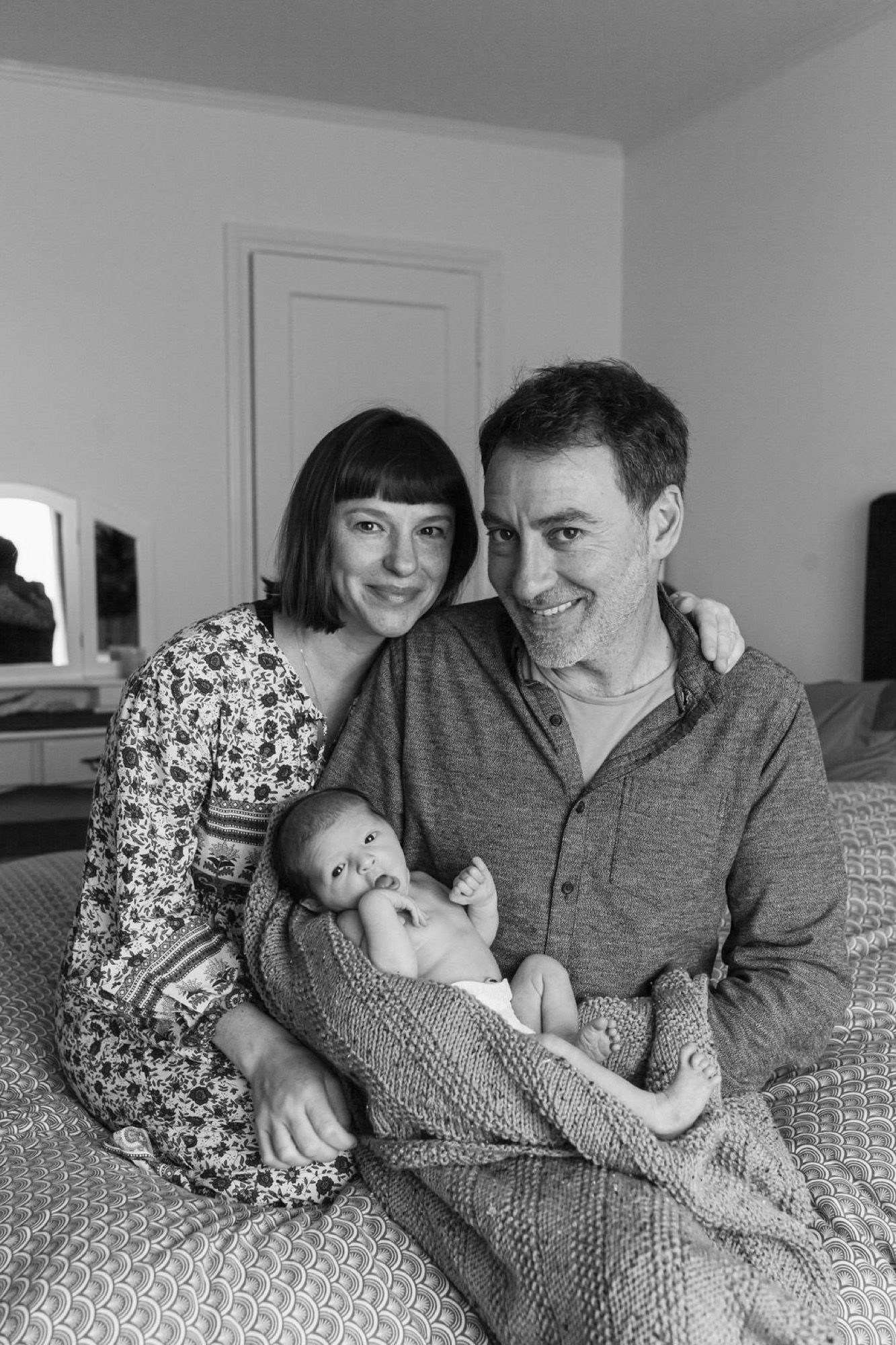 Parents sit together on a bed holding their newborn, both smiling warmly at the camera in soft natural light