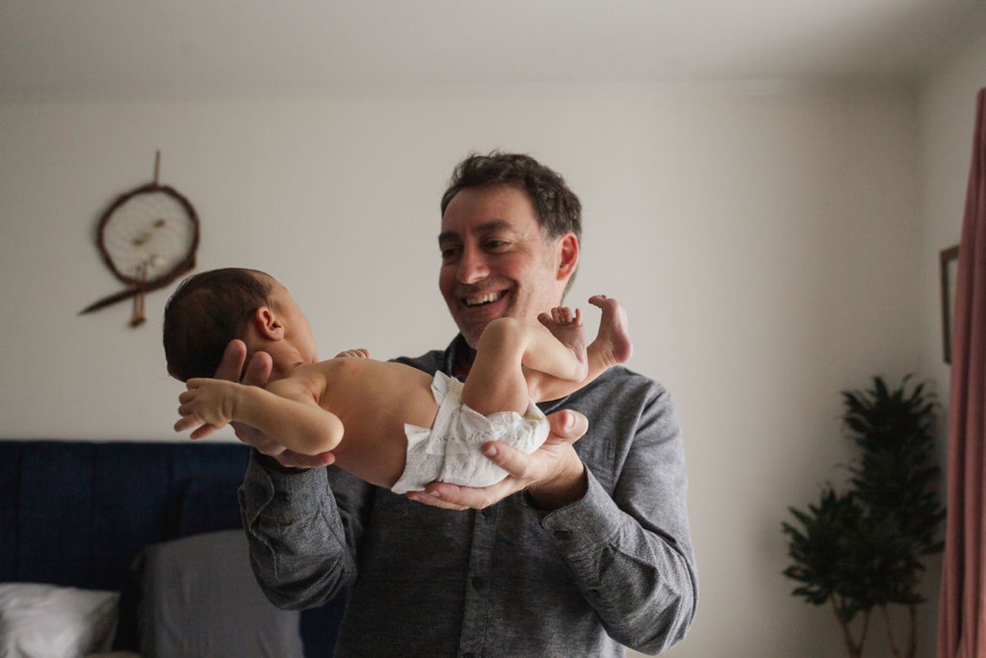 A laughing father holds his infant son aloft in a bright bedroom, the baby's arms stretched wide as morning light floods the intimate moment