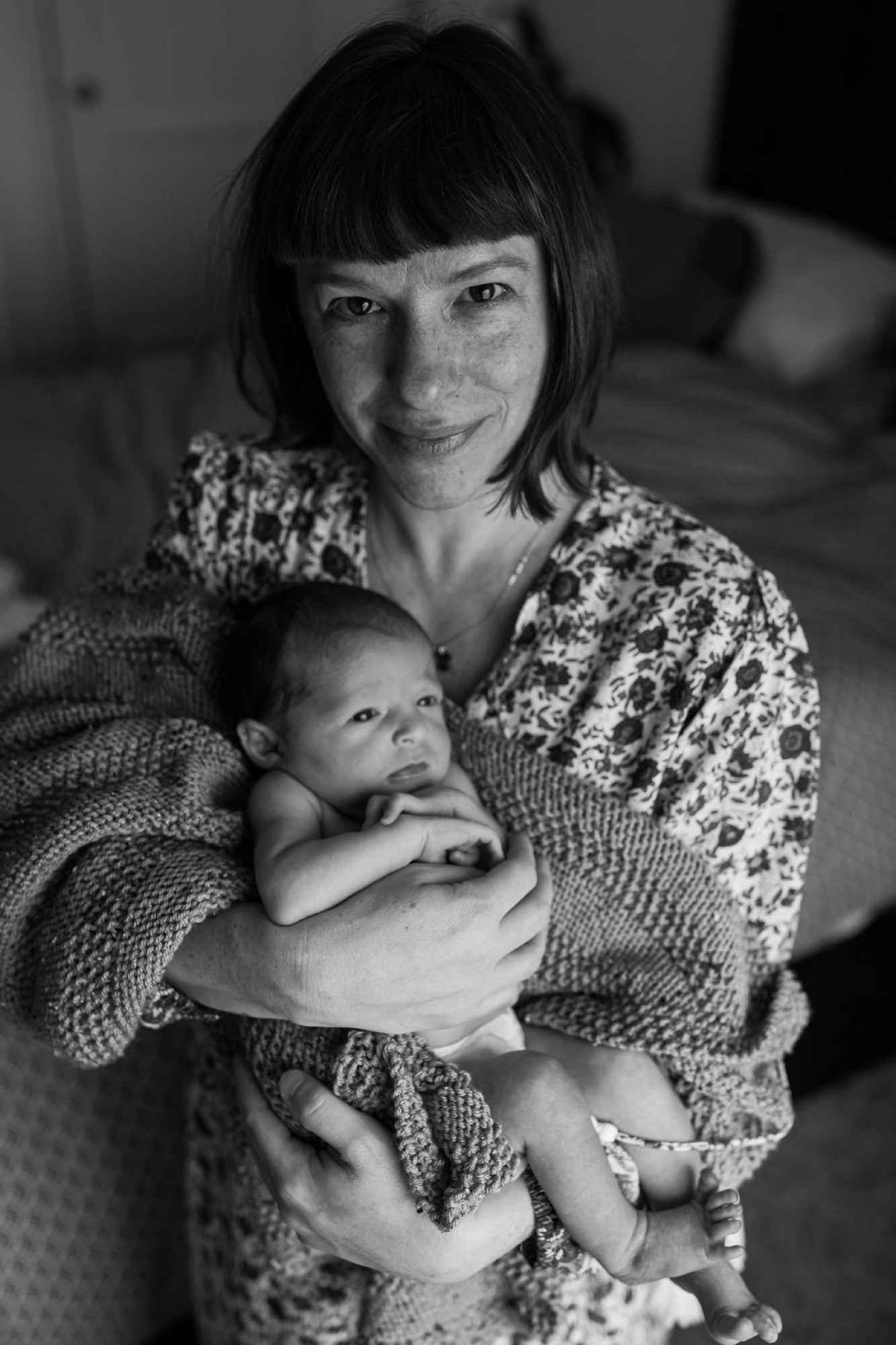 Mother gazes softly downward while cradling her newborn in a textured knit wrap, natural window light creating gentle shadows across their faces