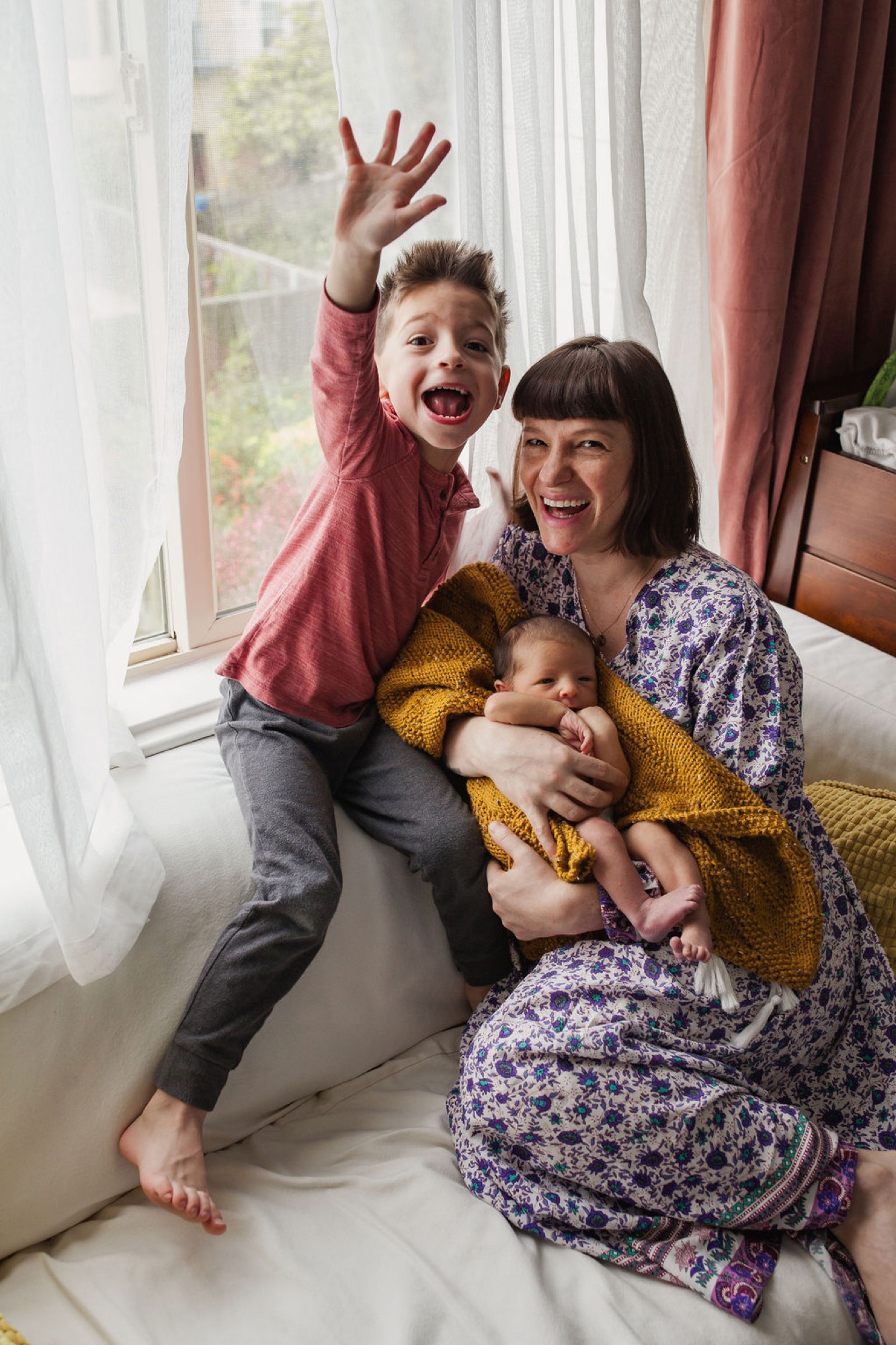A mother sits by a bright window with her two children, the older boy raising his arm joyfully while she holds an infant wrapped in mustard yarn