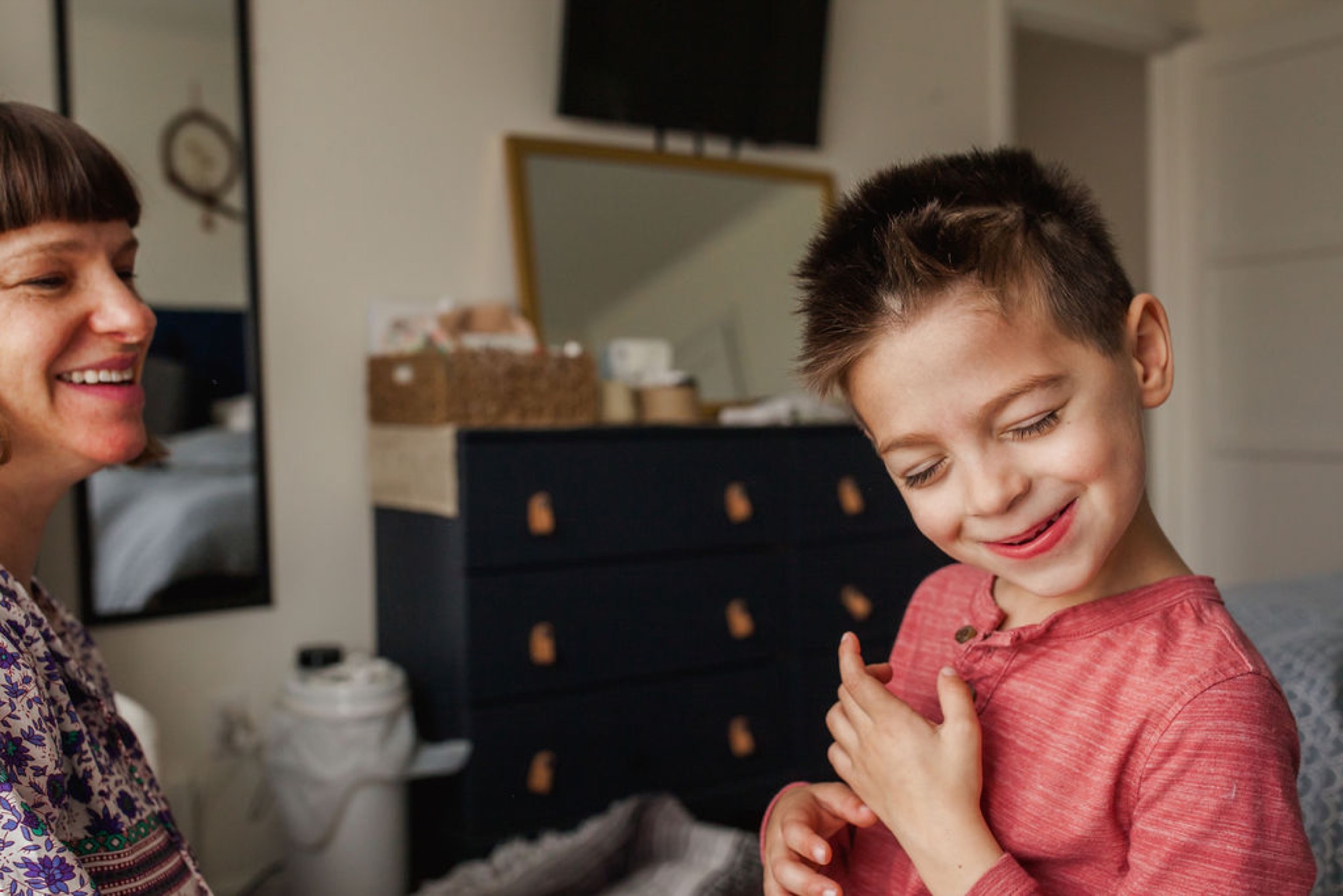 A woman and young boy laugh together in a bedroom, the child wearing a coral pink shirt with genuine delight on his face
