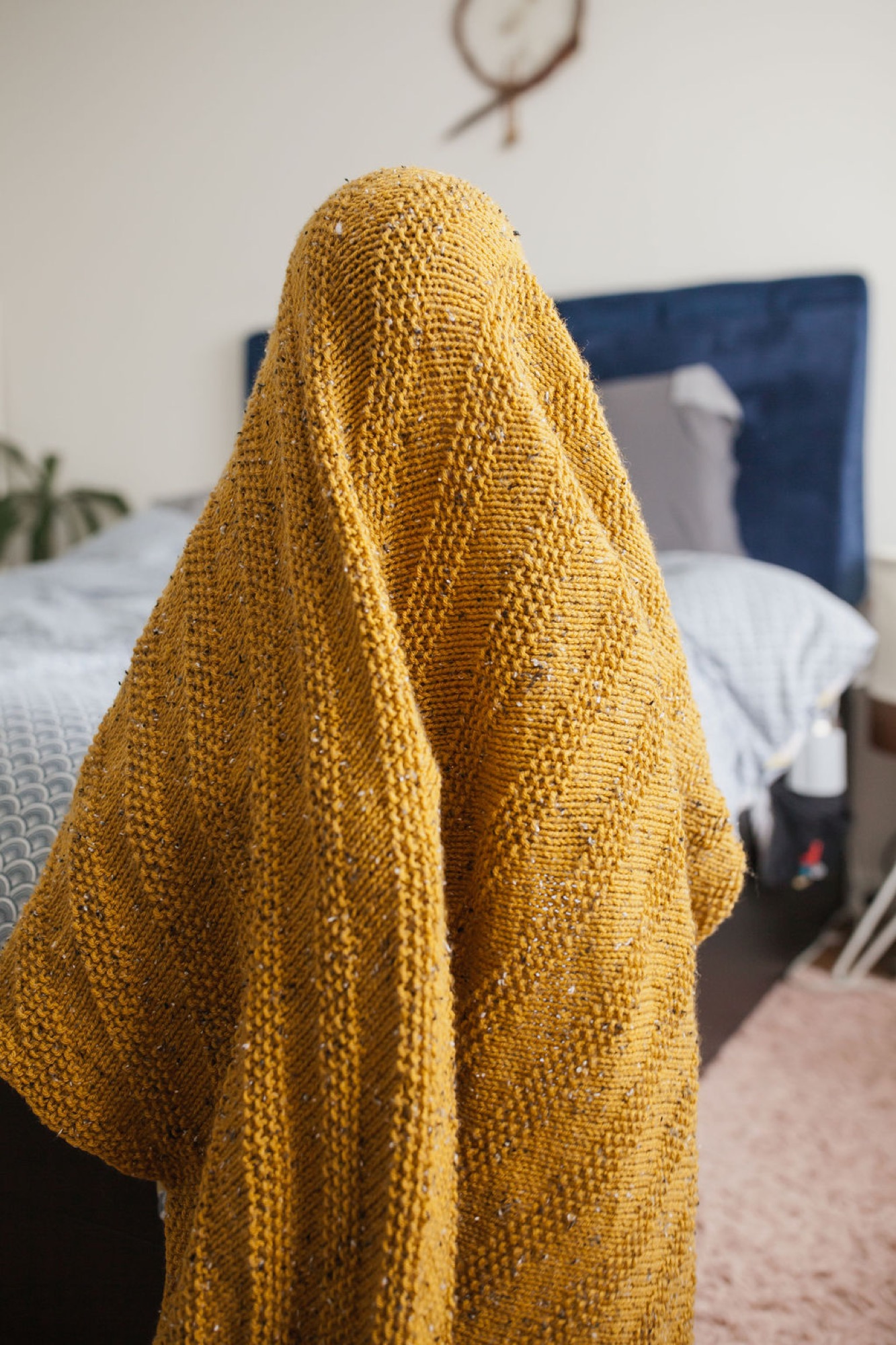 A golden knit blanket drapes over a person's shoulders in a sunlit bedroom, casting soft textured shadows against white bedding