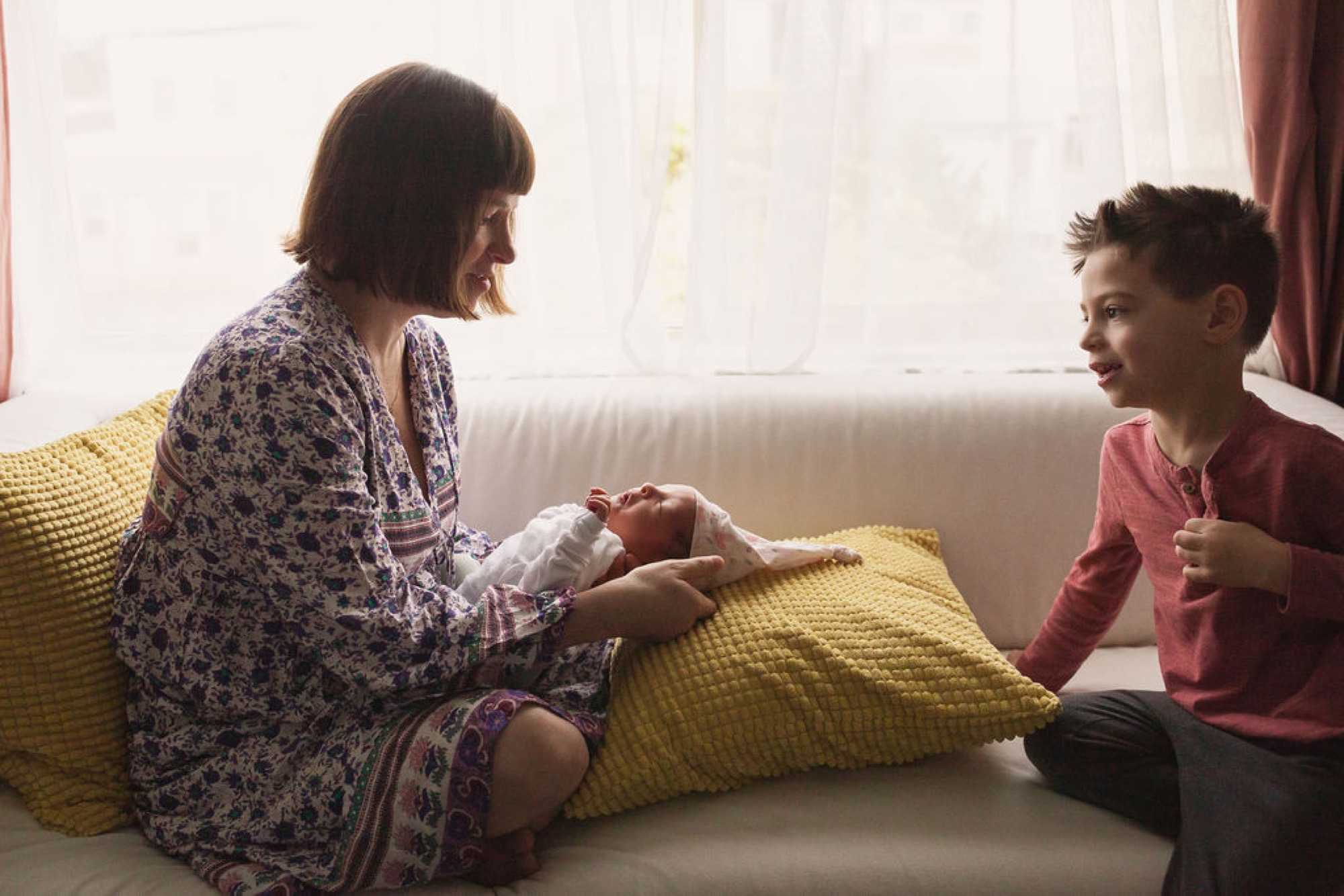 Mother and young son sit together on a bright yellow pillow-laden sofa, sharing an engaged moment while holding a swaddled newborn between them