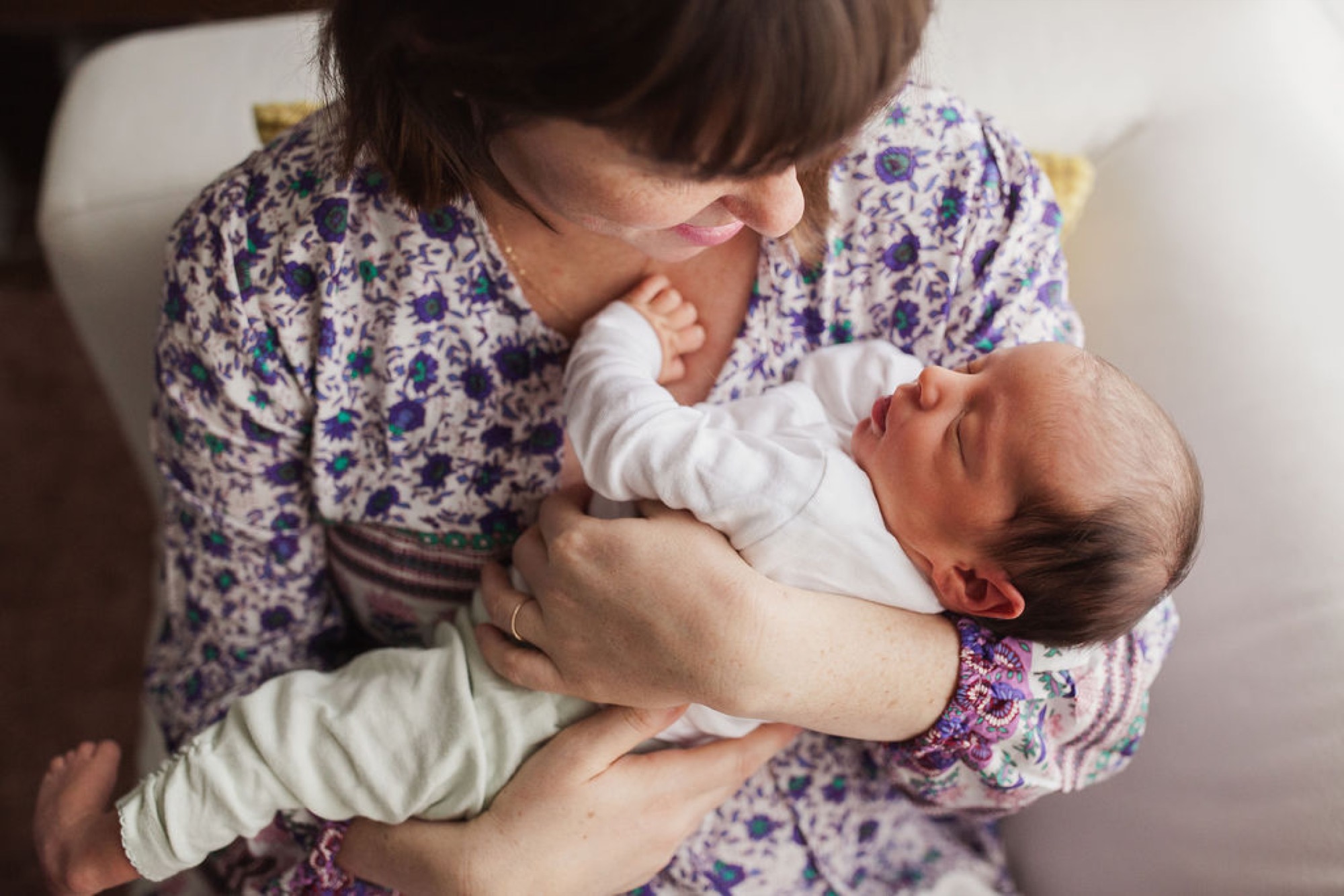 A mother in a blue floral shirt gazes down tenderly at her newborn wrapped in white, cradling the infant close to her chest