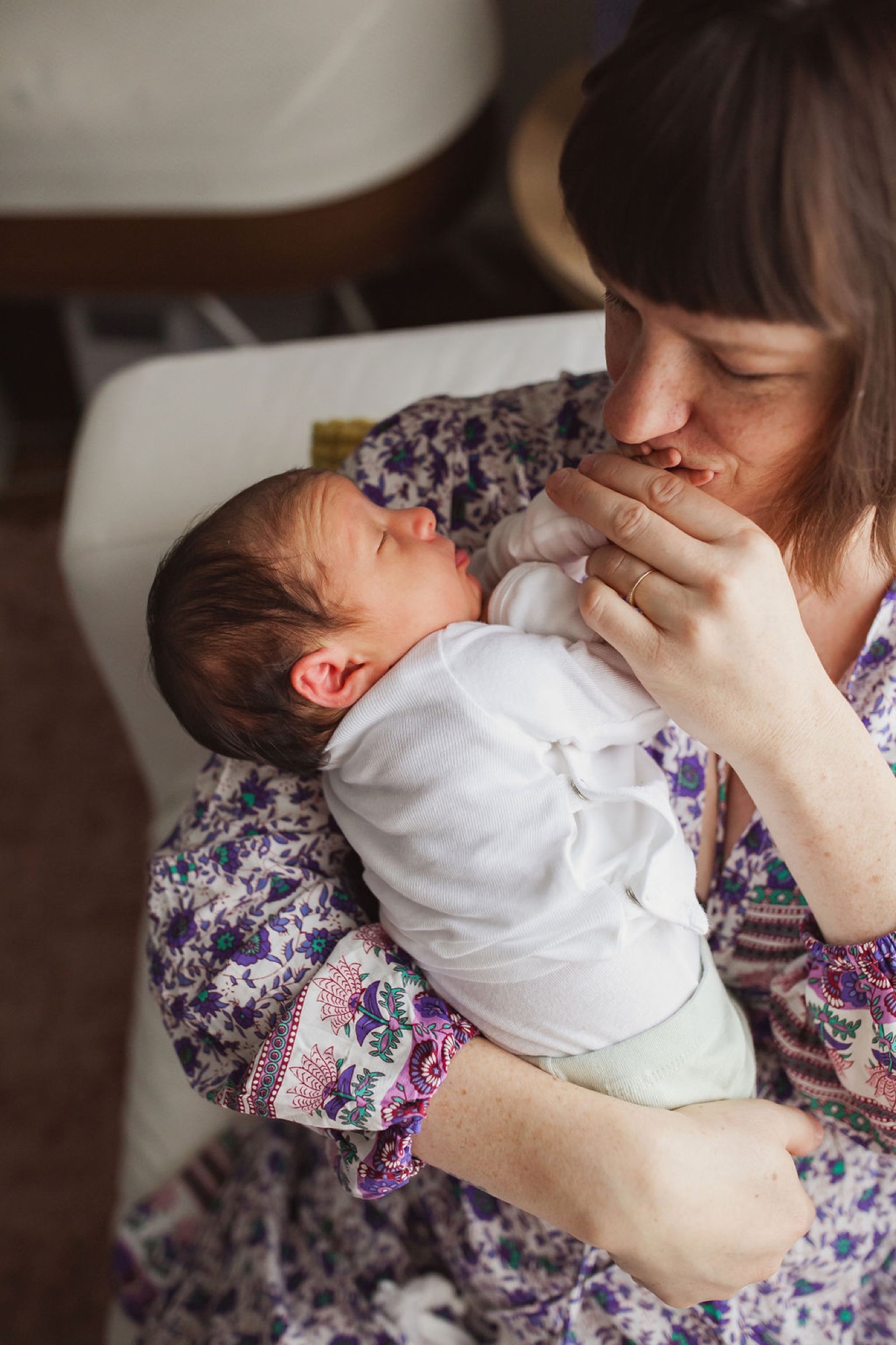 A woman in a purple floral dress tenderly kisses her newborn's forehead while cradling the swaddled infant against her chest in warm indoor light