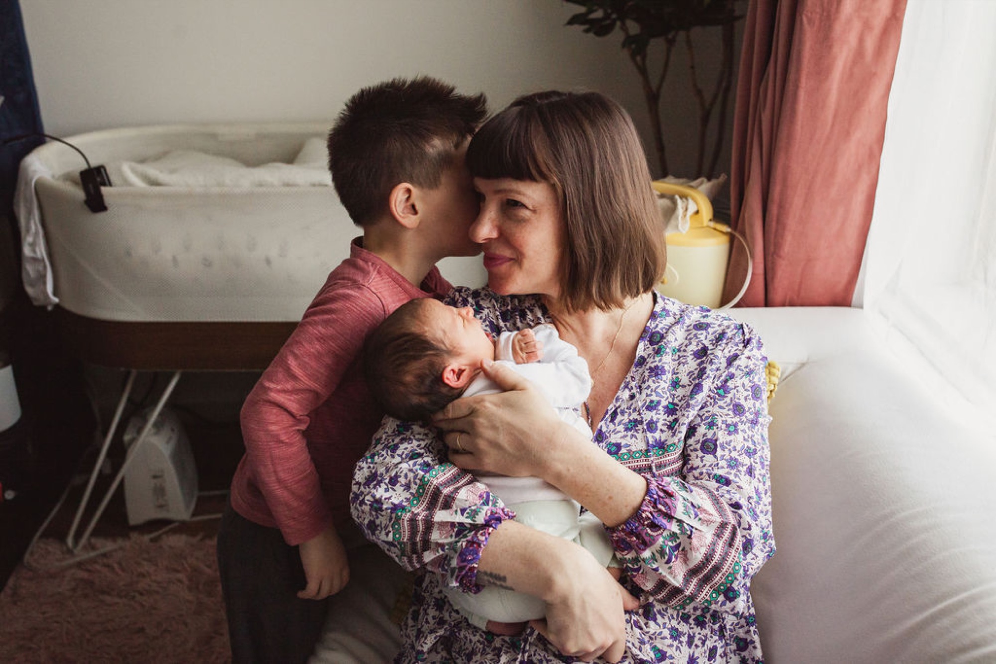 A woman in a floral purple dress holds her newborn while her toddler son leans in for a kiss in a bright bedroom