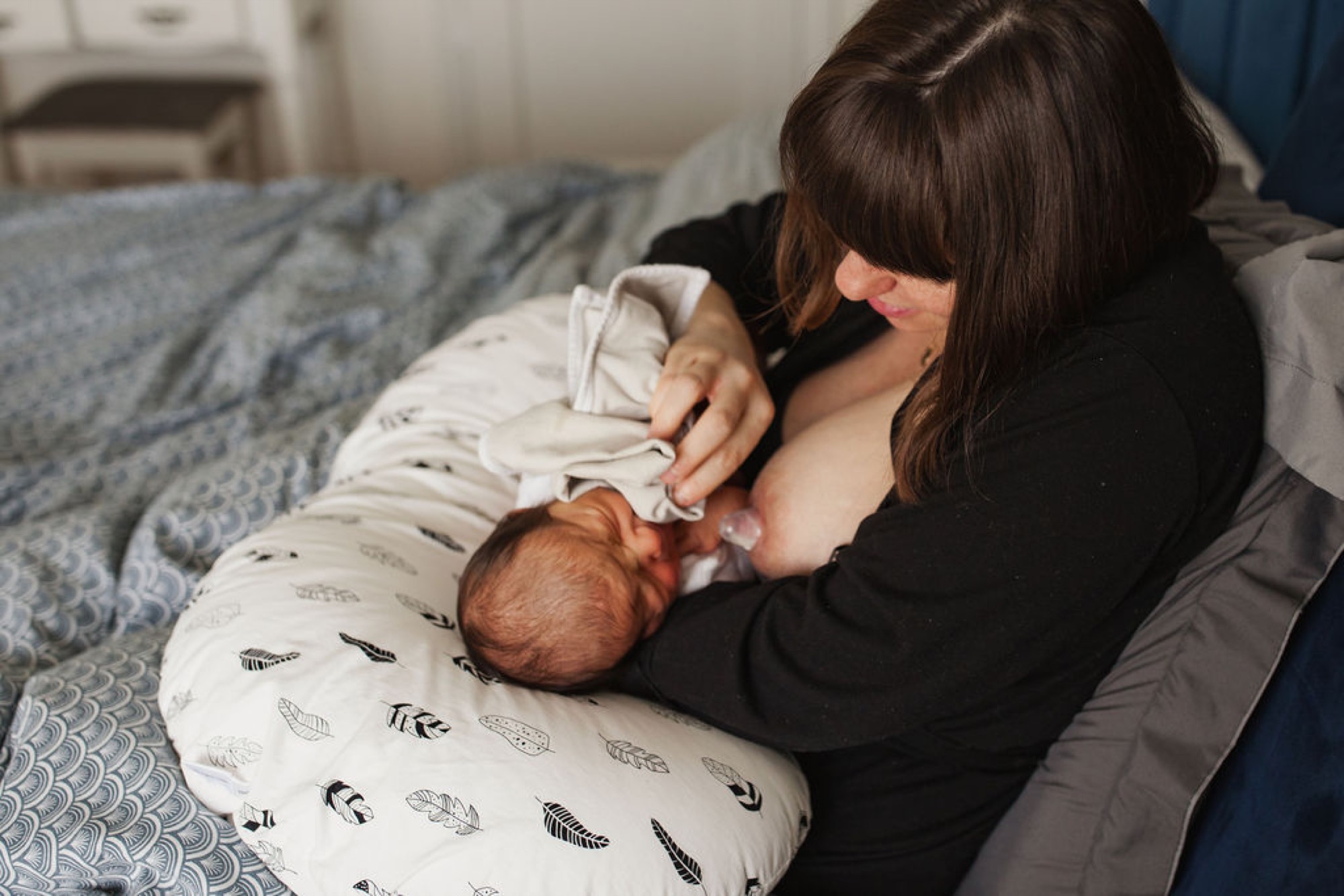 Mother gazes down at newborn nursing while seated on a bed with patterned bedding, soft natural light illuminating their intimate moment