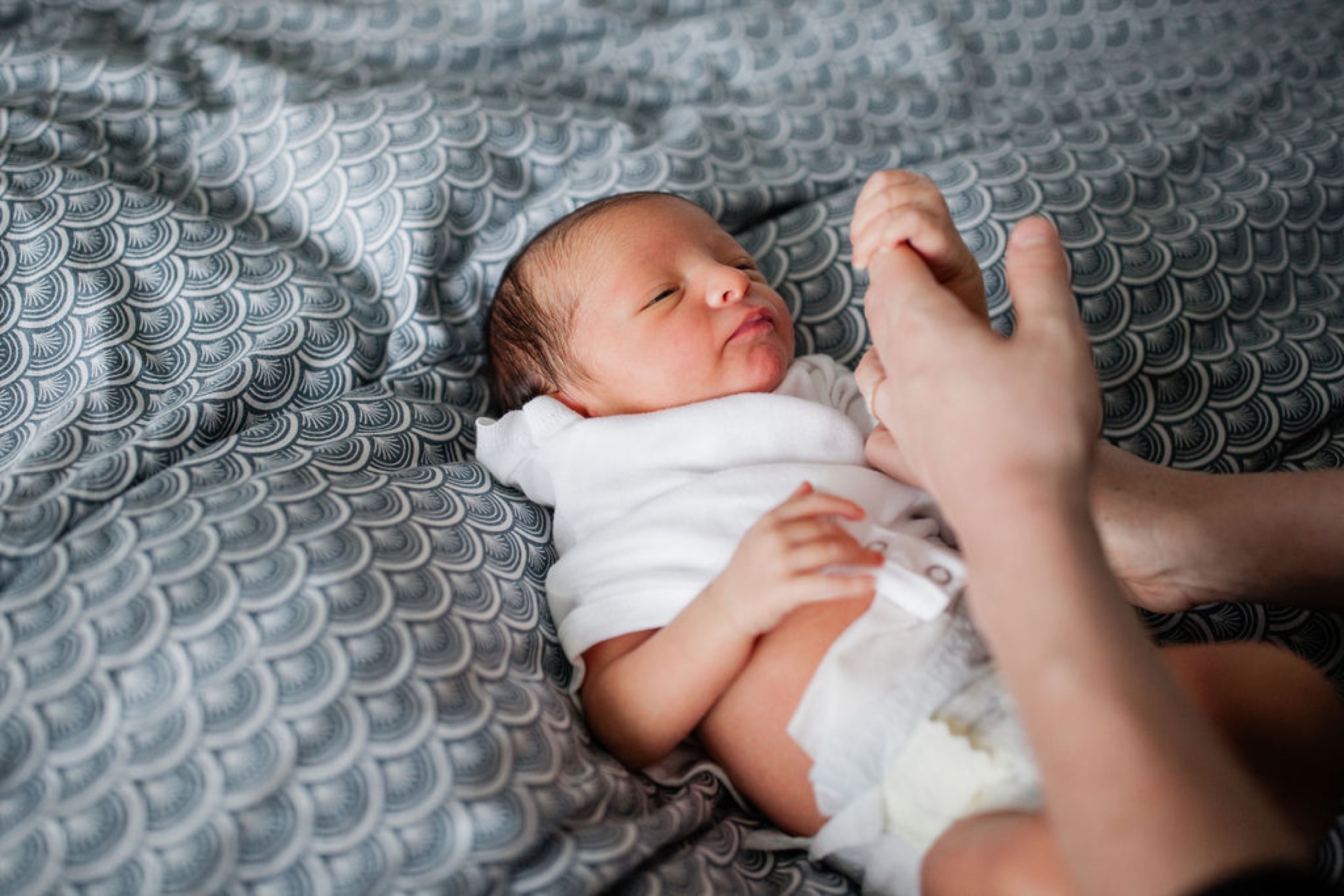 A newborn sleeps peacefully on a gray patterned blanket while an older sibling's hand gently touches their arm in tender connection