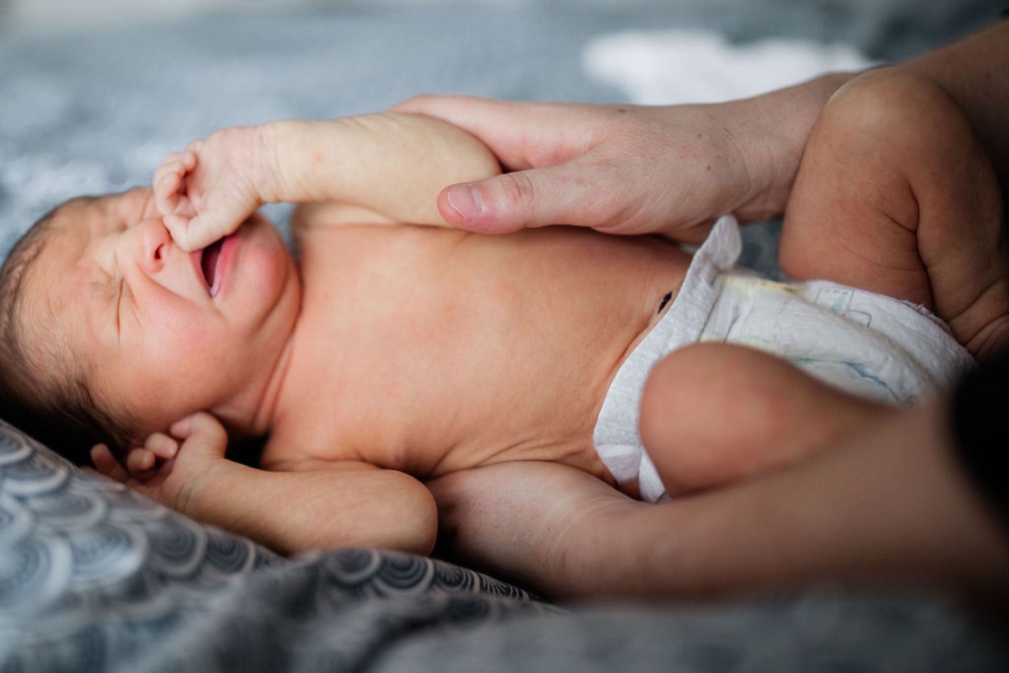 A newborn's tiny hand grasps an adult's finger while lying peacefully in a white diaper on patterned fabric