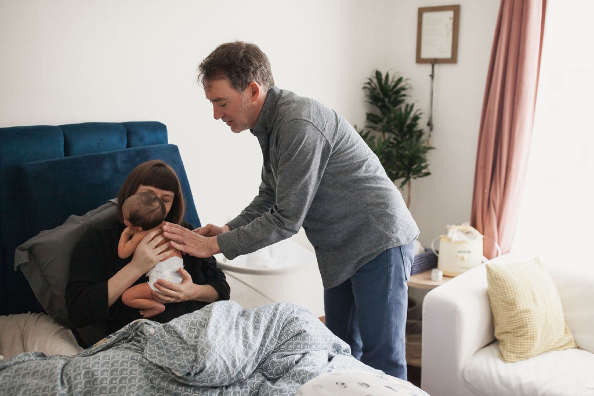 Father leans over mother cradling newborn on bed with blue headboard, soft natural light from window illuminating intimate family moment