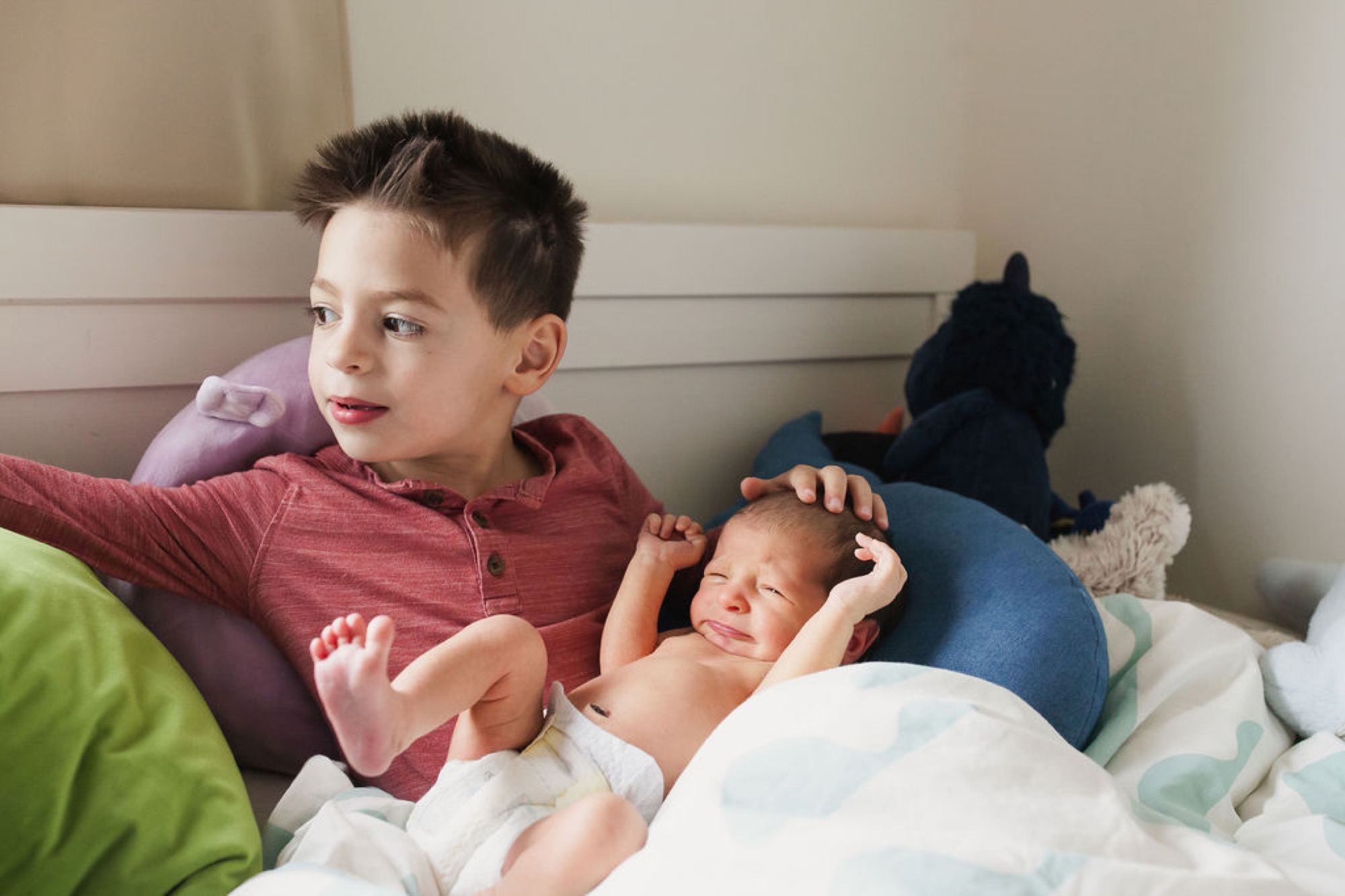 An older brother in a burgundy shirt gently cradles his newborn sibling on a bed, gazing down with tender affection while the baby sleeps peacefully