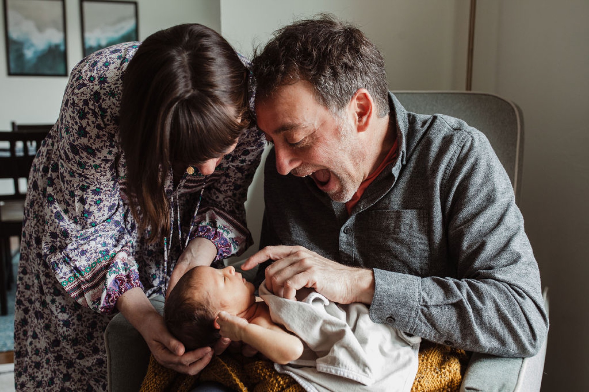 Parents lean in together over their newborn, foreheads nearly touching as they gaze down with tender smiles in soft natural light