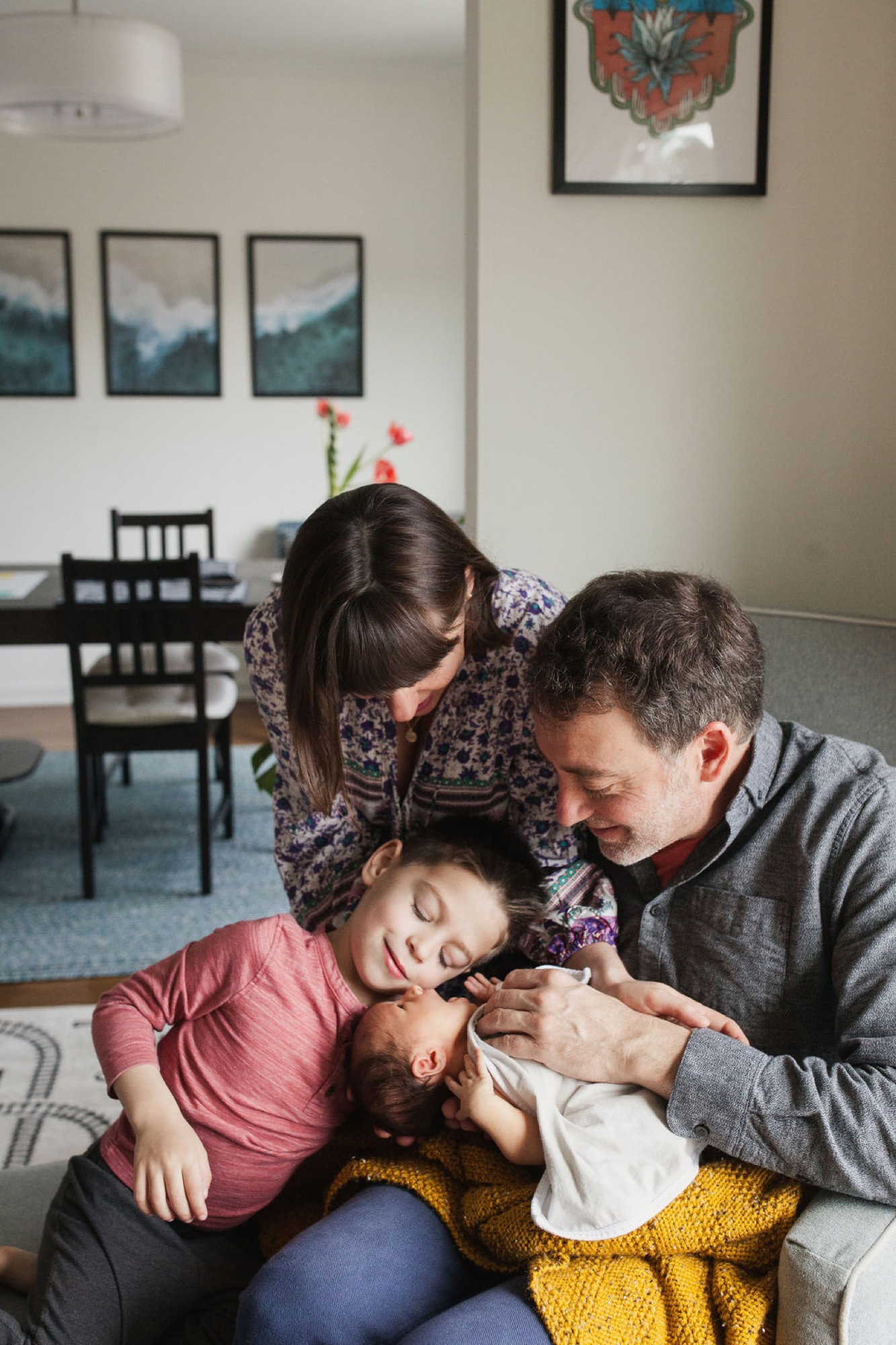 Family of four sits closely on a gray couch, parents cradling their newborn while an older sibling leans in with a gentle smile, warm natural light filling the modern living room