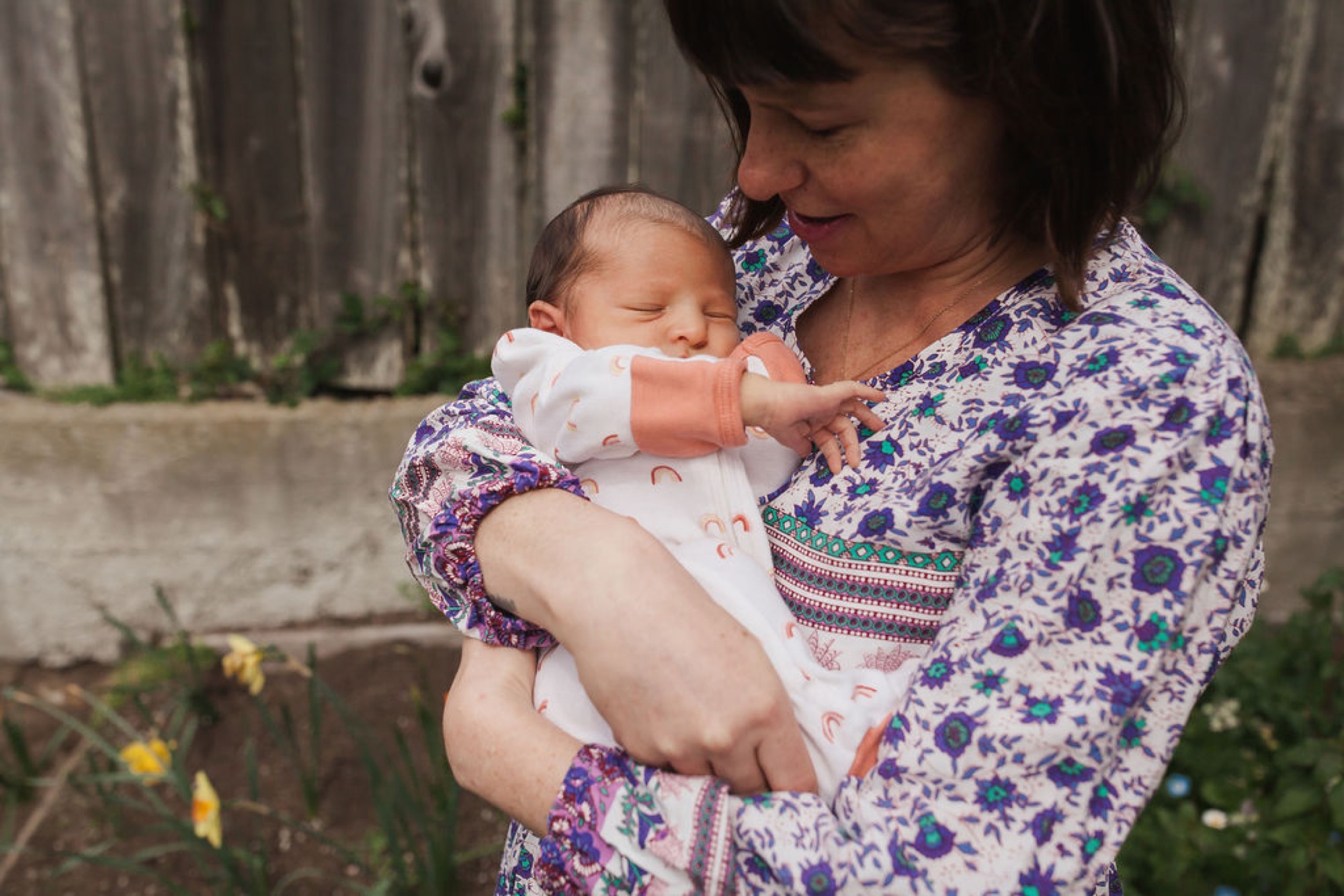 Mother in a blue floral cardigan gazes down at her peacefully sleeping newborn wrapped in white, positioned against a weathered wooden fence