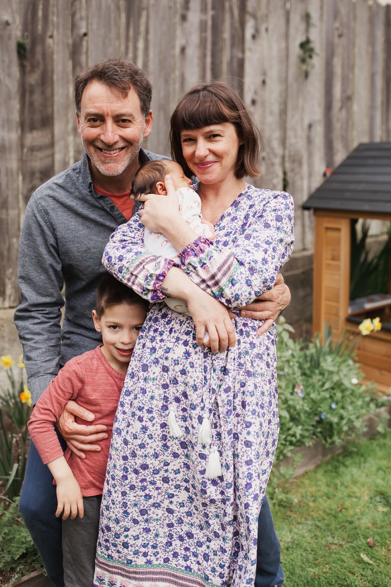 Family of four smiles warmly in their backyard, with father's arm around mother holding newborn while older son stands in front against wooden fence