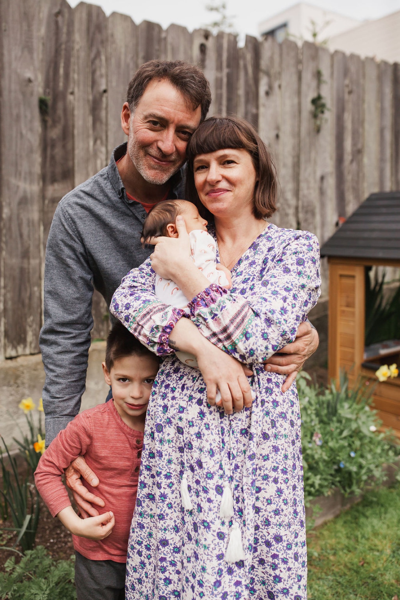 Family of four poses together in their backyard, with father and mother smiling warmly while holding their newborn and young son in a casual embrace