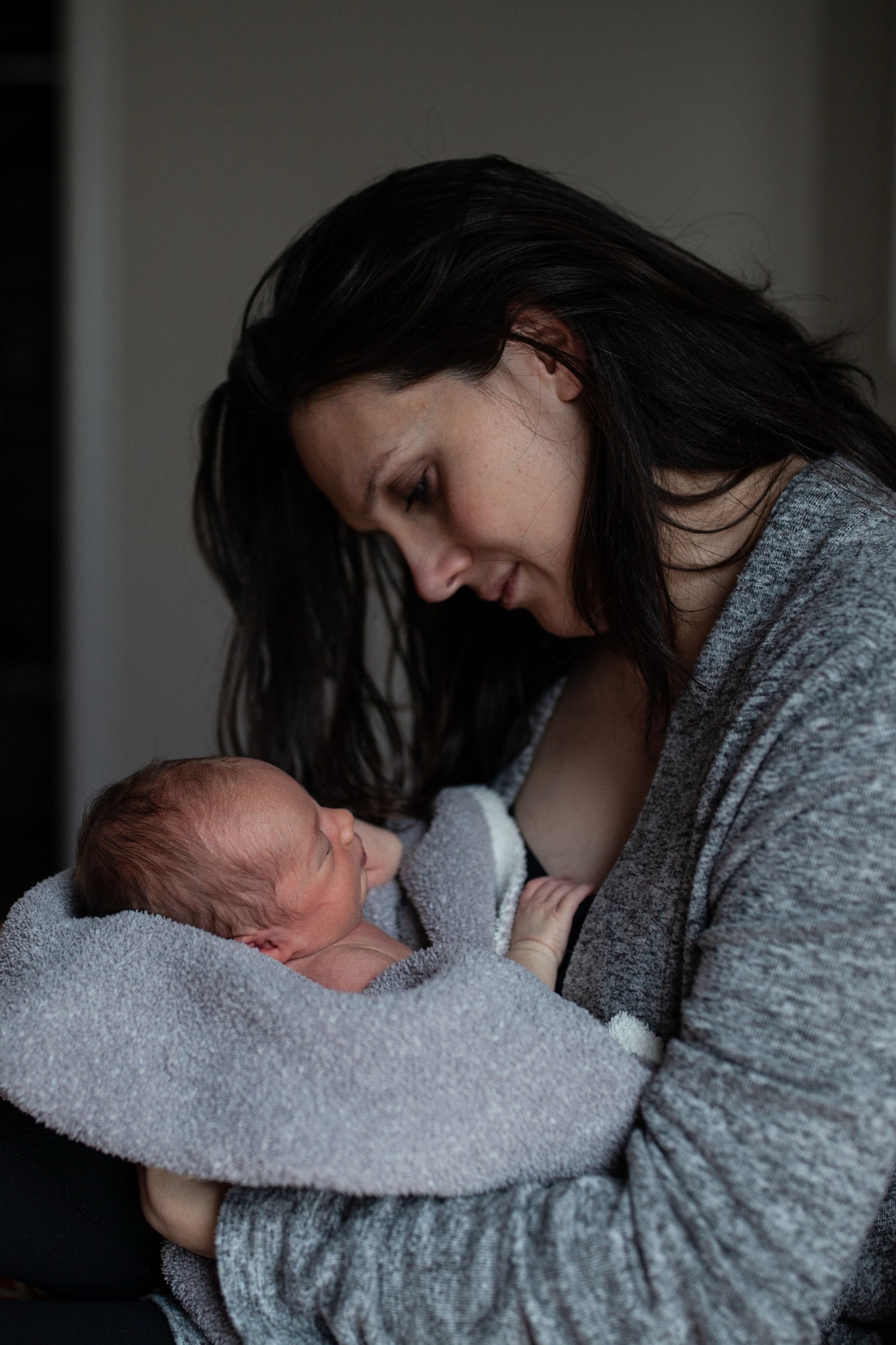 Mother gazes down tenderly at newborn cradled in her arms, soft window light illuminating their intimate moment