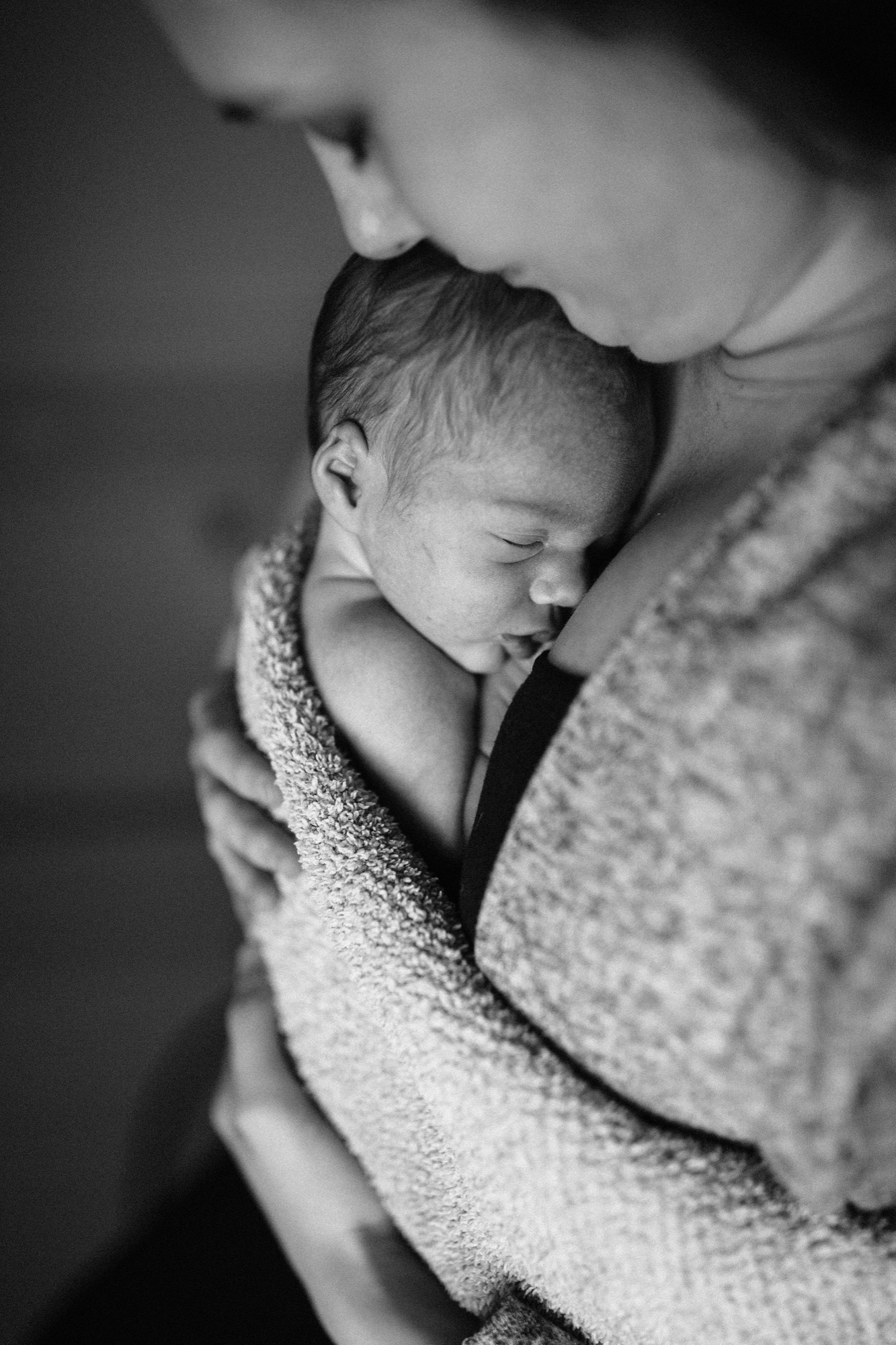 A parent cradles a sleeping newborn wrapped in a soft knit blanket, their face tender and close in intimate black and white light