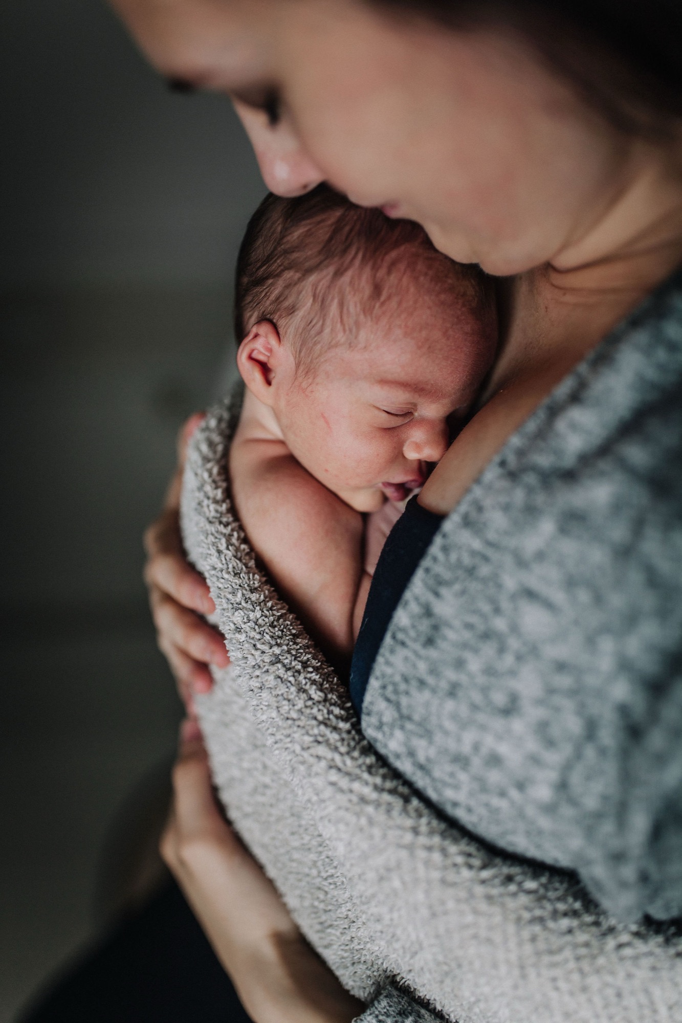 Mother gazes down at her newborn wrapped in soft gray fabric, capturing tender intimacy and peaceful contentment