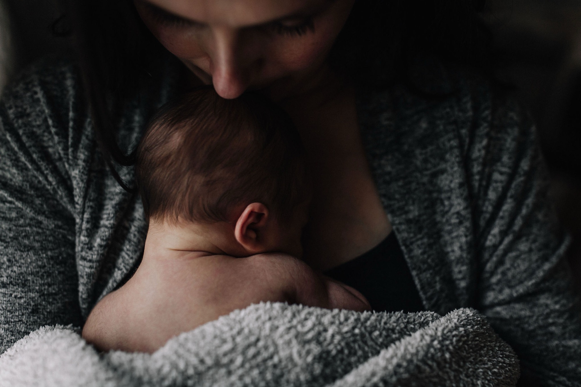 A parent cradles their newborn close, tenderly kissing the baby's head in soft, intimate home lighting