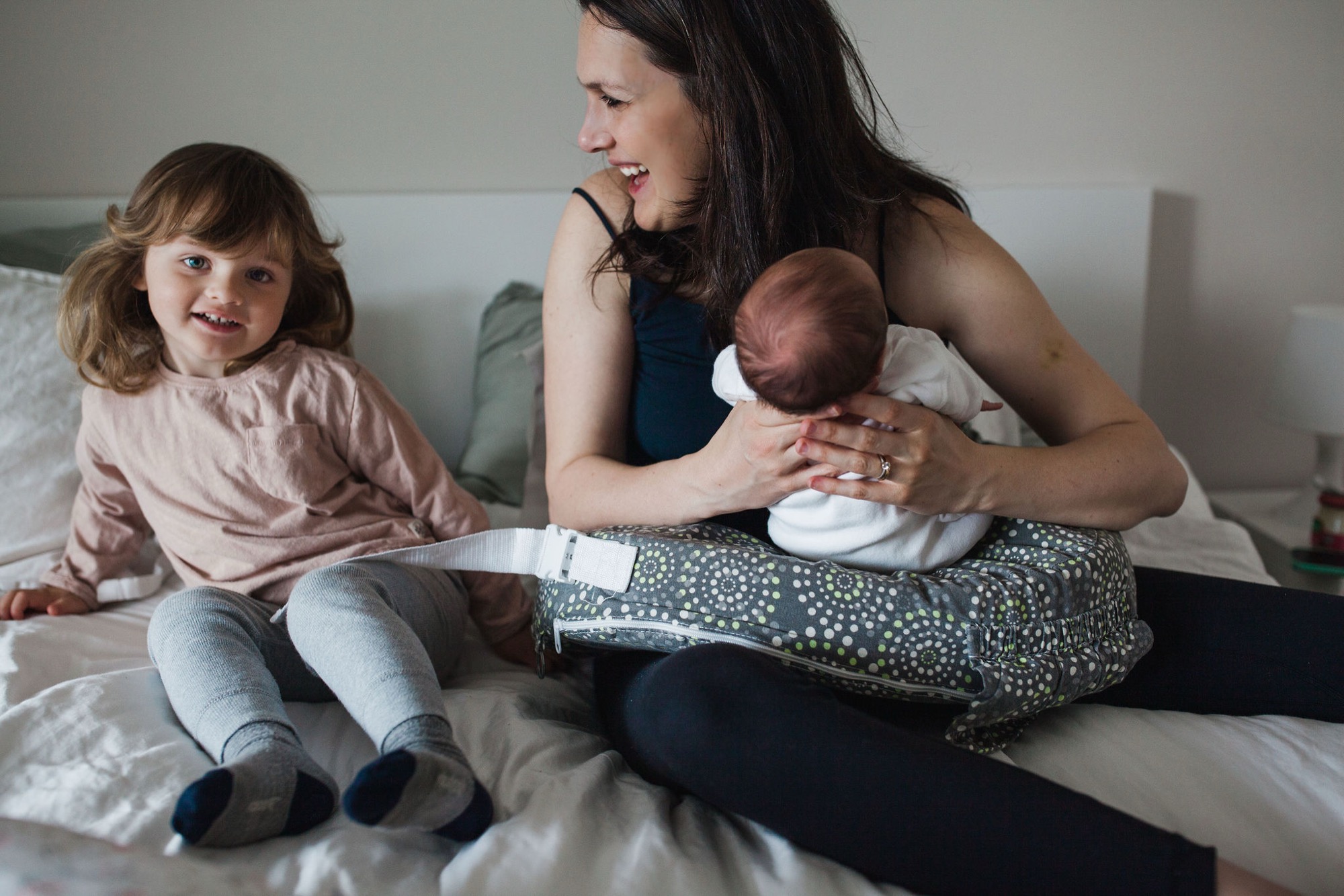 A mother sits on a bed with her newborn and toddler daughter, smiling warmly while the older sibling gazes at the camera with gentle curiosity
