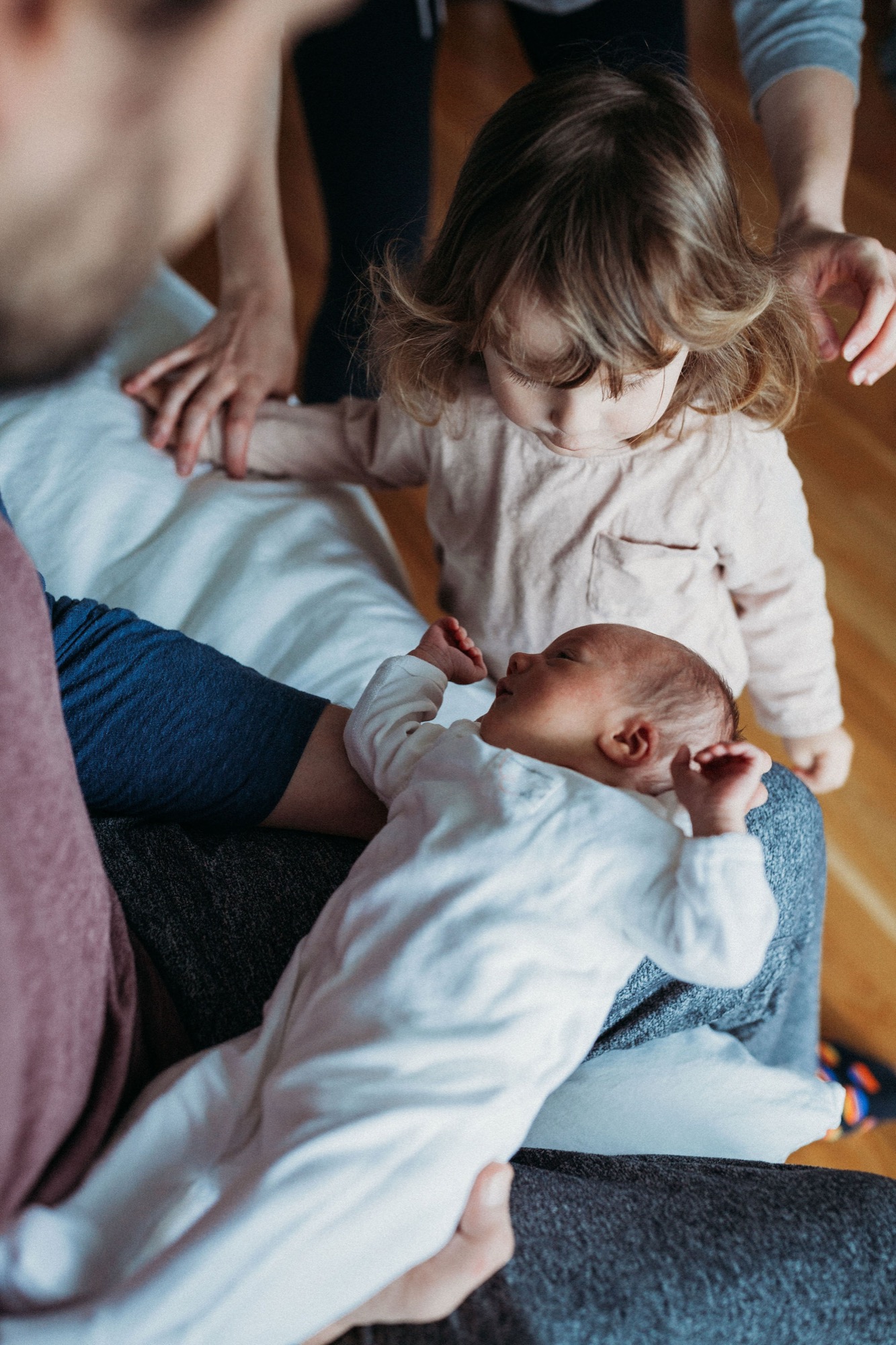 Toddler reaches toward newborn sibling cradled in parent's lap during intimate home session