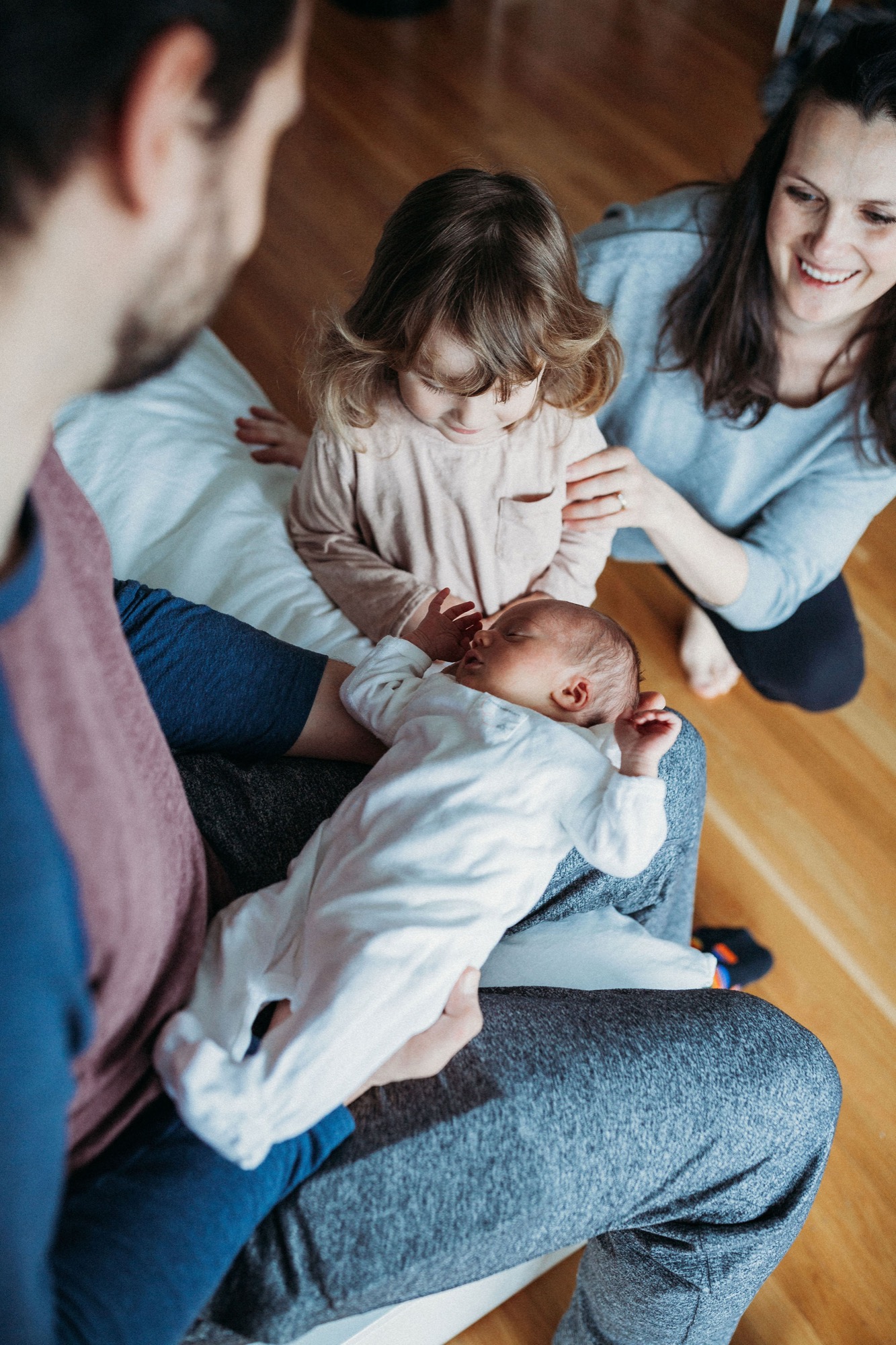 Young family sits on wooden floor, parents cradling newborn while toddler reaches toward baby with genuine curiosity and joy