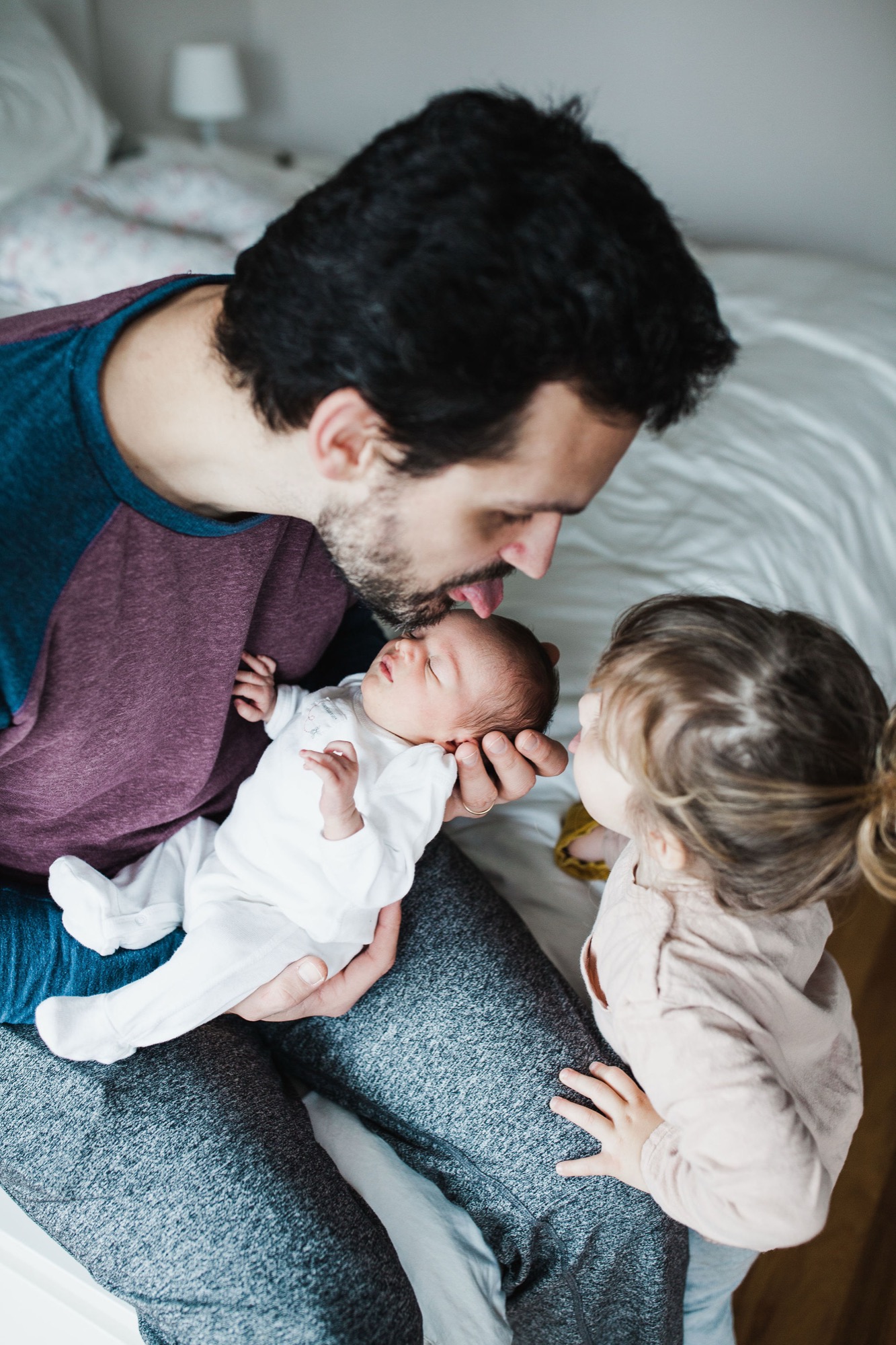 Father gazes tenderly at newborn while older sibling leans in close on white bedding, captured in soft natural light