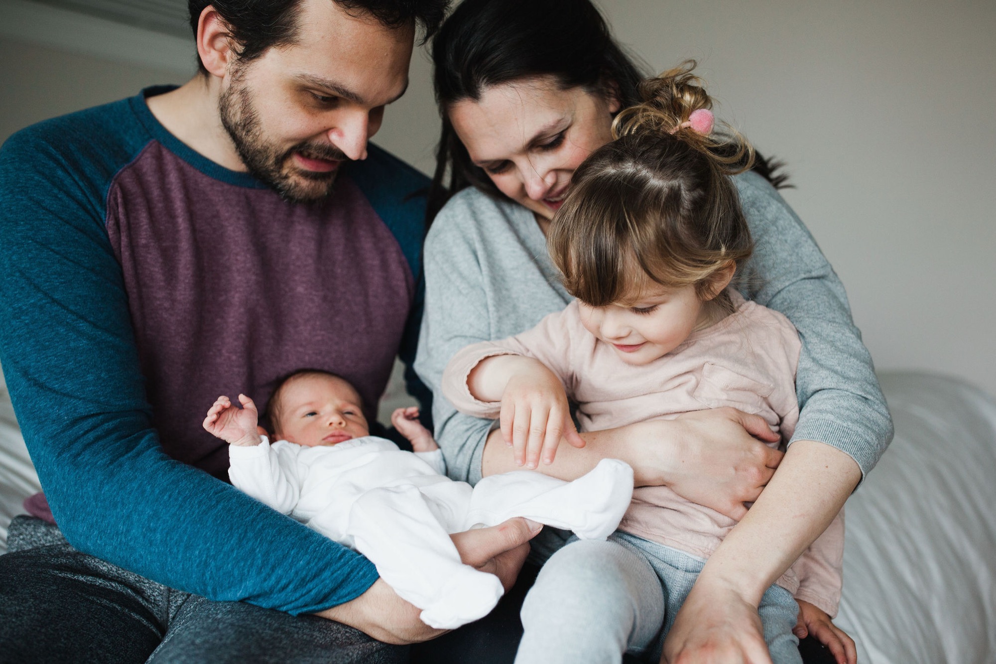 Parents and toddler lean close around newborn on bed, soft natural light illuminating genuine smiles and tender family connection