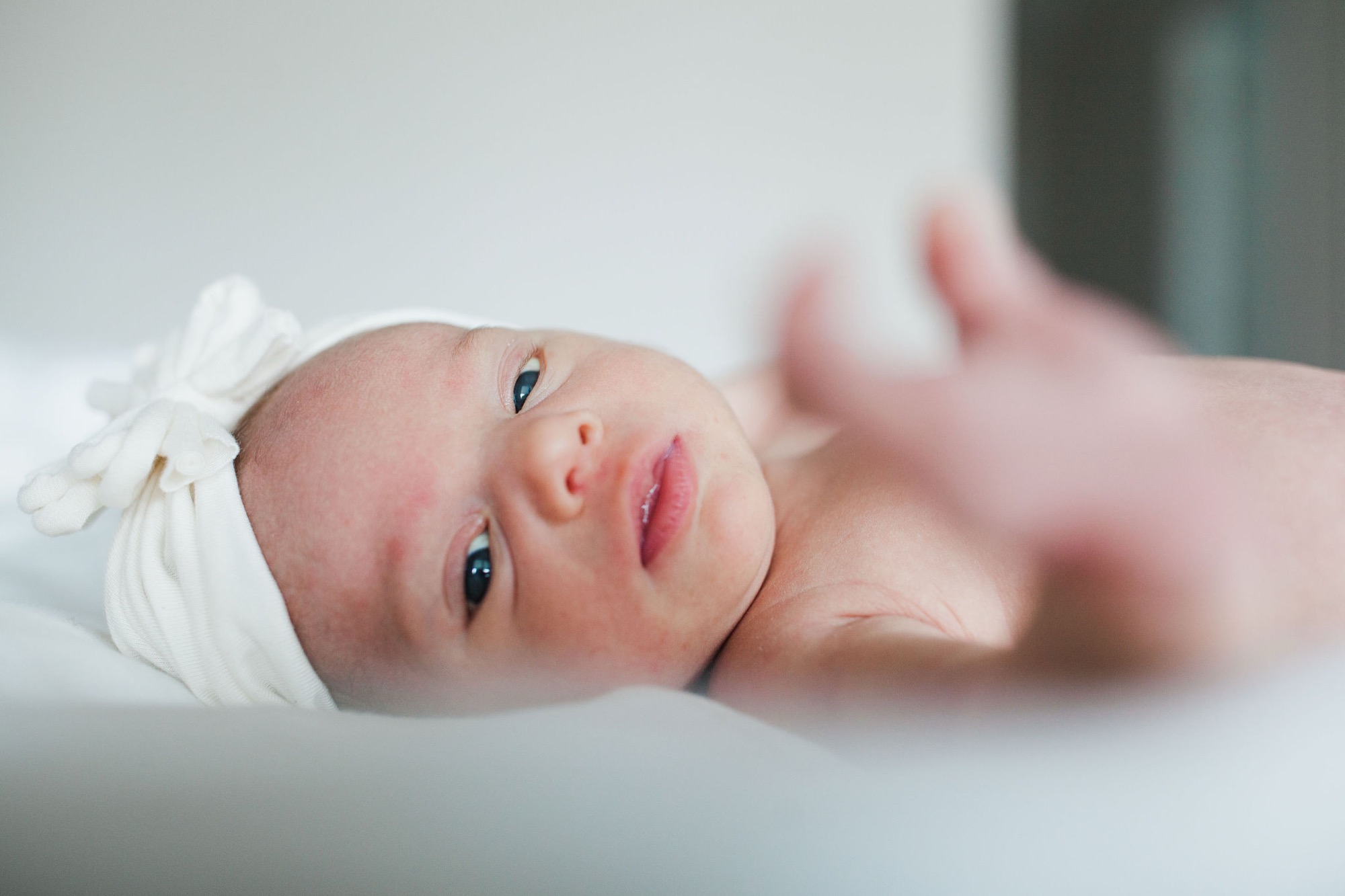 Newborn lies swaddled in white fabric, dark eyes alert and gazing directly at camera with soft, peaceful expression