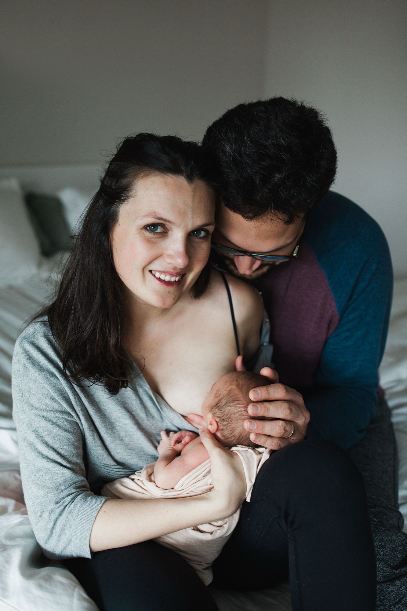 A beaming mother holds her newborn while her husband embraces them both from behind on a soft bed, natural window light illuminating their genuine joy