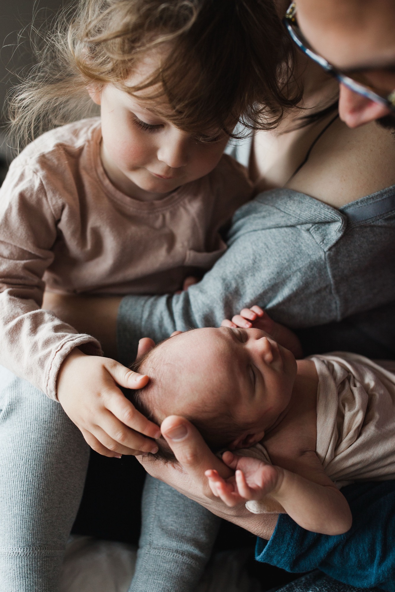 Older sibling gently cradles newborn's head while mother holds them both, soft natural light illuminating their tender connection