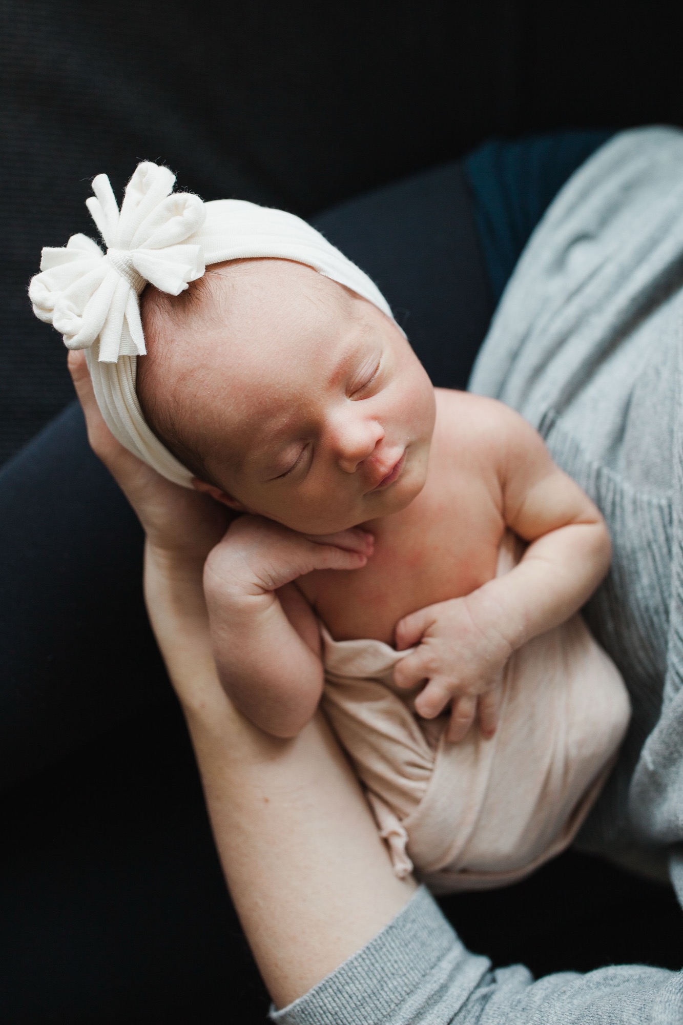 Sleeping newborn with white bow headband nestled in parent's cradling hands against dark background