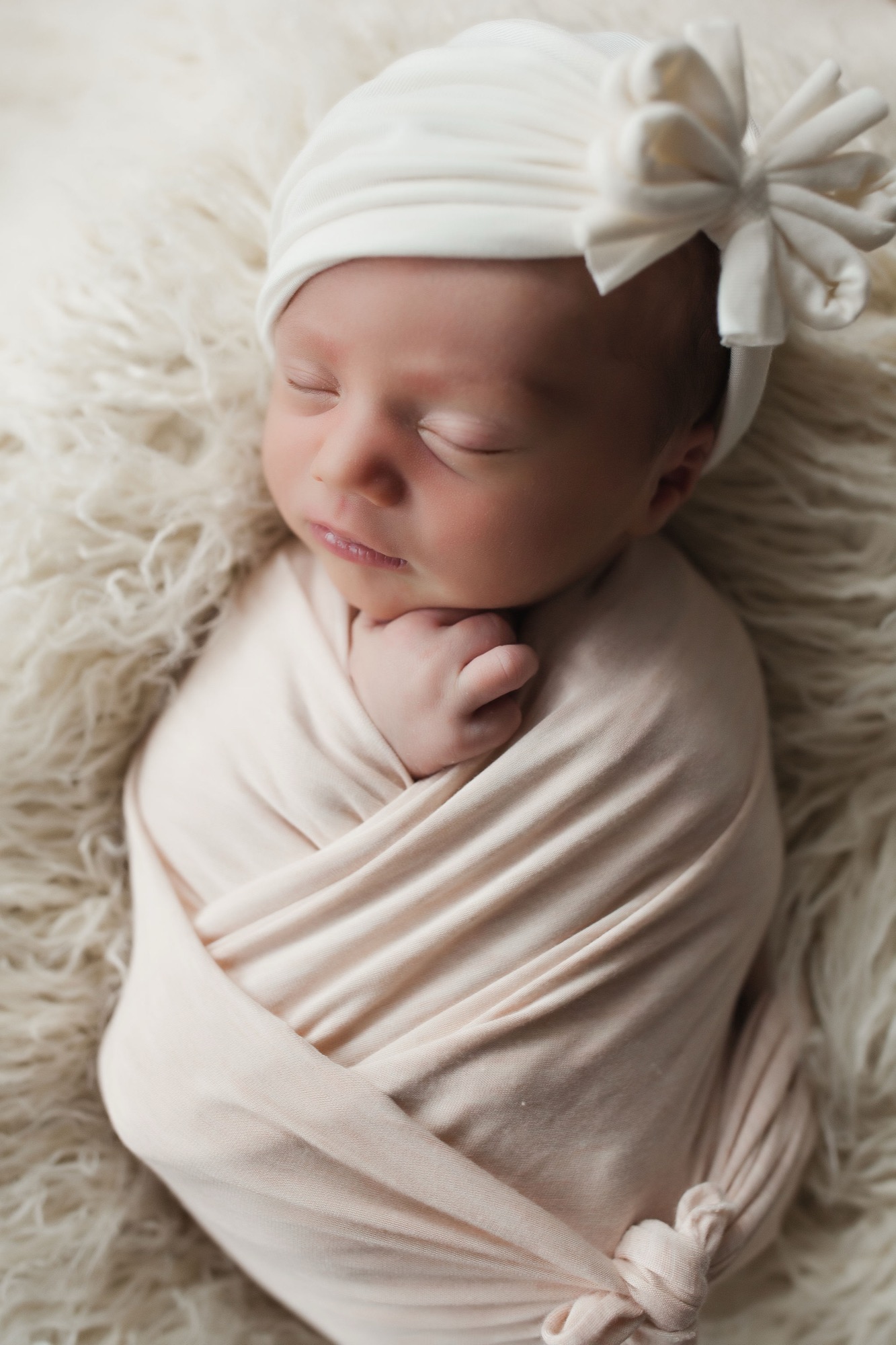Peacefully swaddled newborn in cream fabric with white bonnet, resting on textured neutral bedding with serene expression