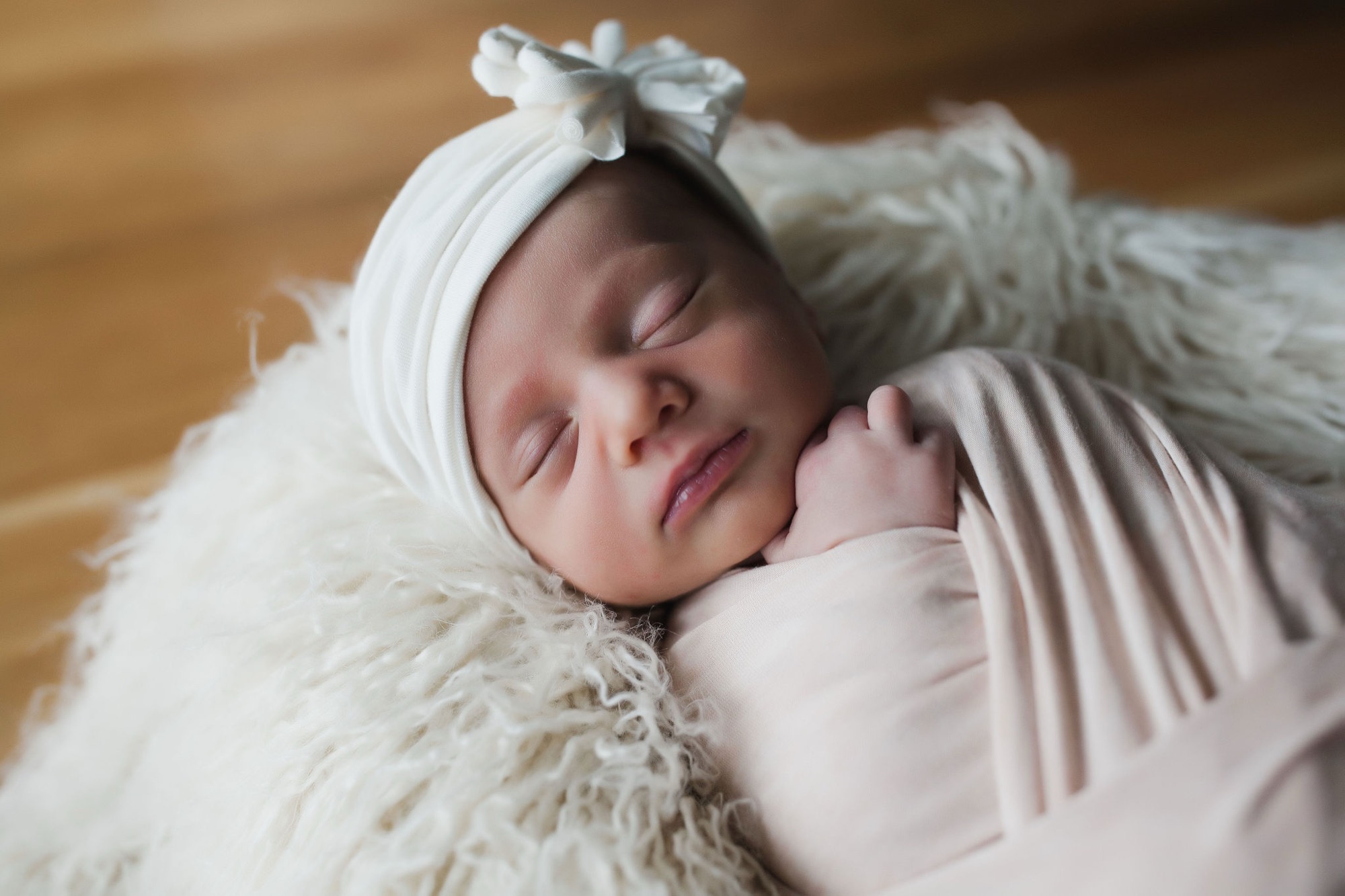 Sleeping newborn swaddled in cream fabric with a delicate bow headband, resting peacefully on soft textured bedding