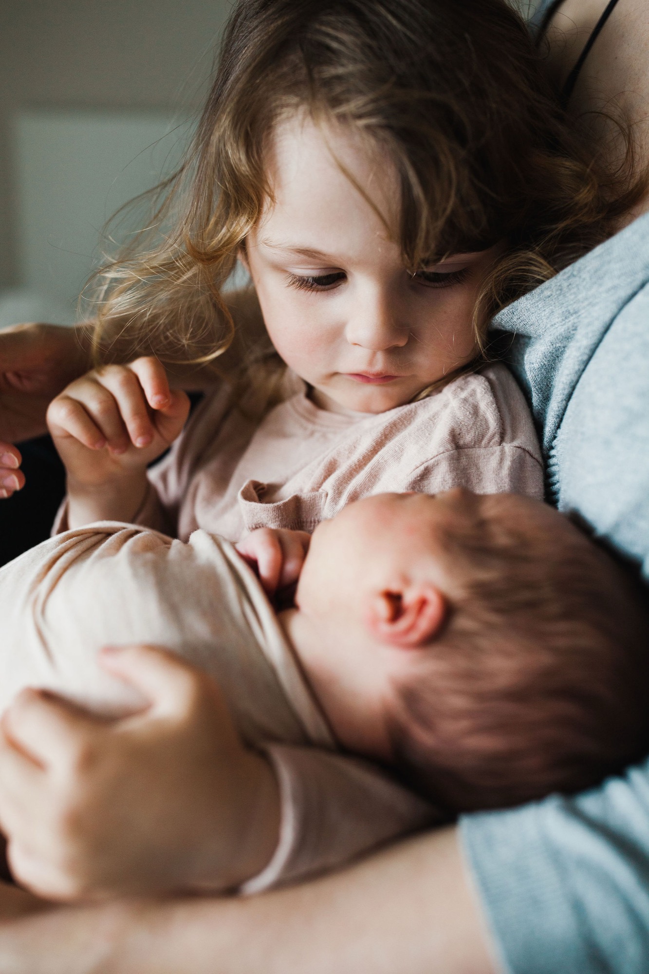 Toddler gazes tenderly at newborn sibling cradled in parent's arms, natural window light illuminating their gentle connection