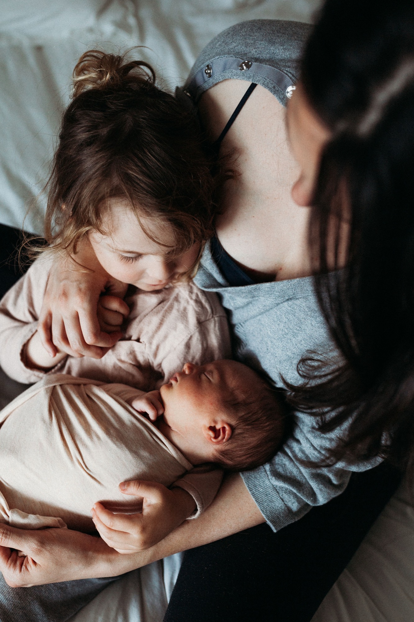 A mother gazes down at her newborn cradled against her chest while an older toddler nestles close, bathed in soft natural light