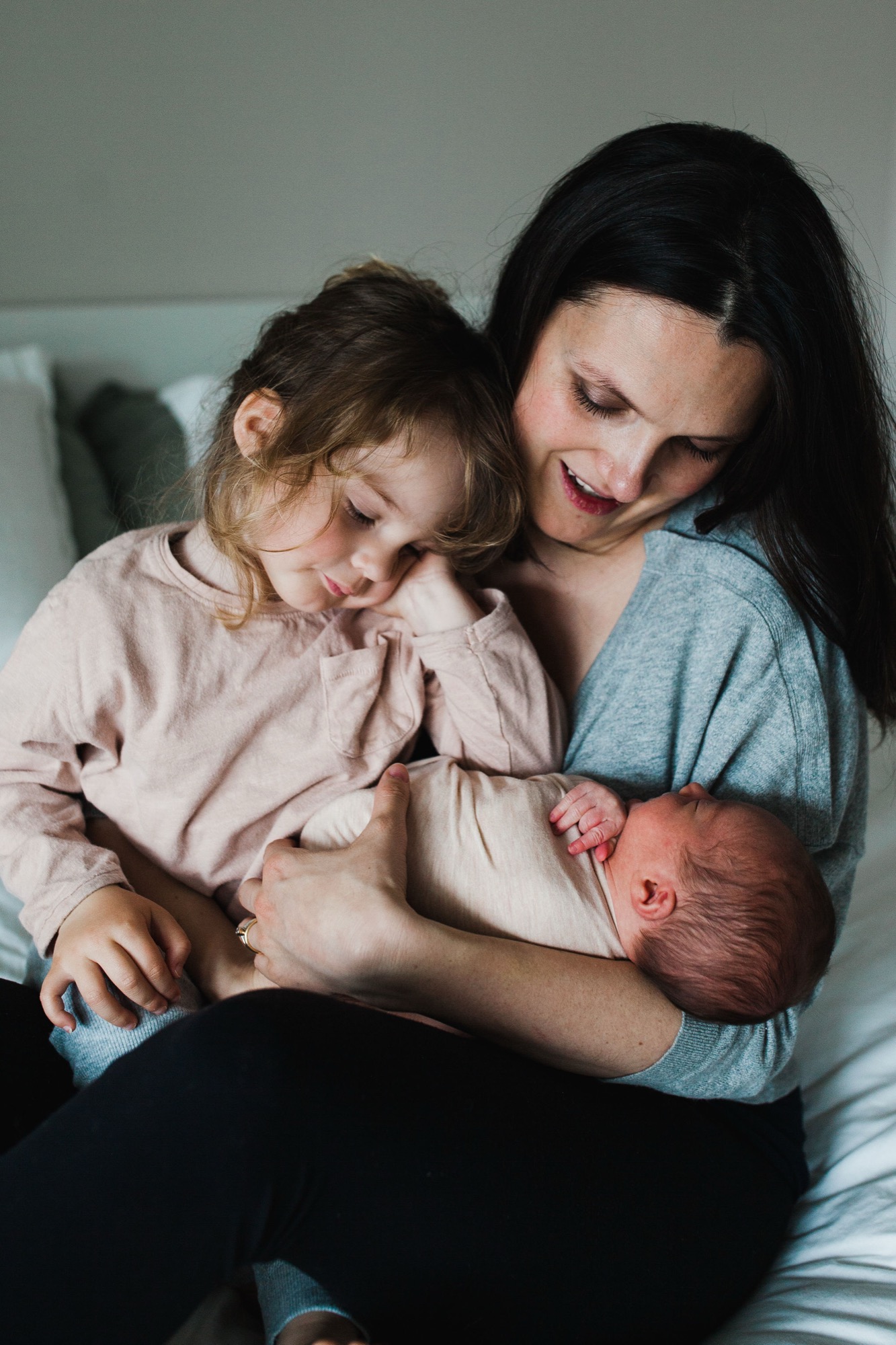 Mother holds newborn and toddler on white bedding, all three gazing downward with genuine warmth and connection