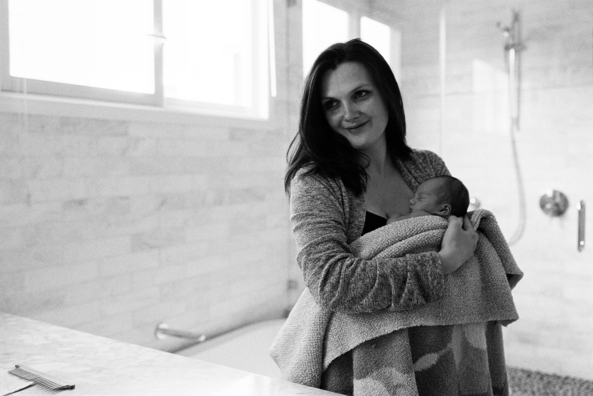 A mother glances back warmly while cradling her swaddled newborn in a sunlit bathroom, soft film grain in the quiet morning light