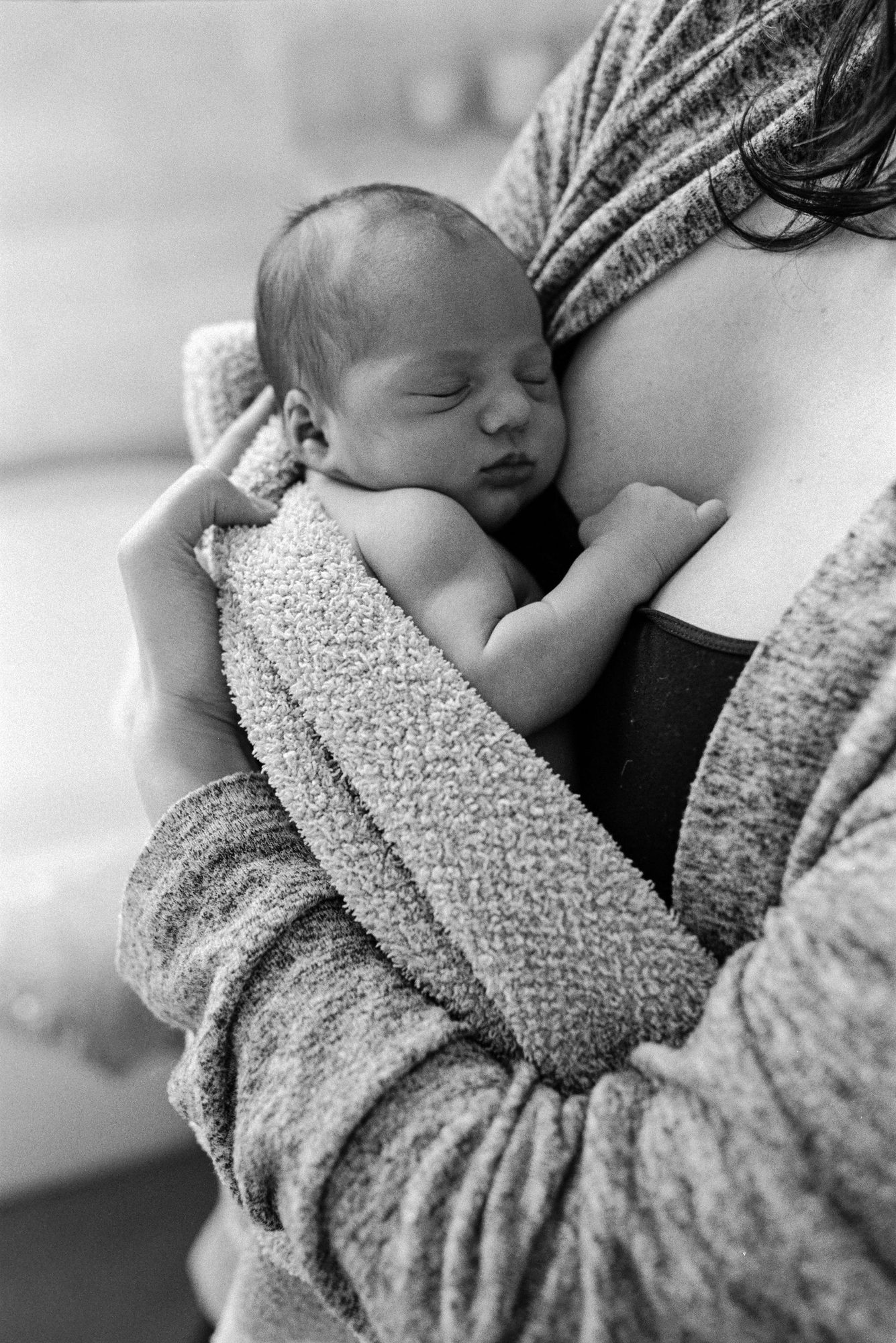 Newborn sleeps peacefully against mother's chest, tiny hand curled near her collarbone in an intimate black and white close-up