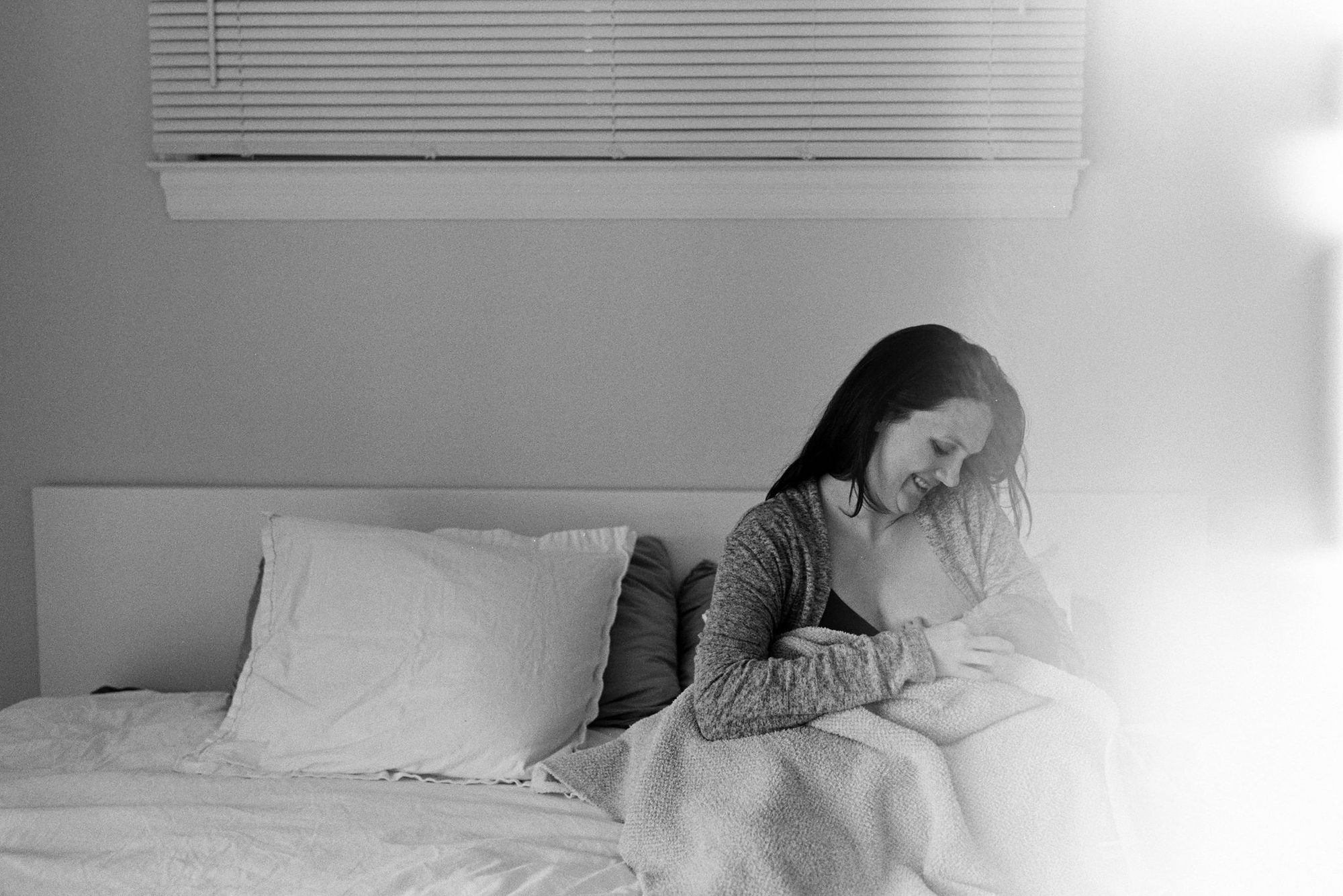Mother sits on the edge of the bed cradling her swaddled newborn, dramatic window light flaring softly across the room