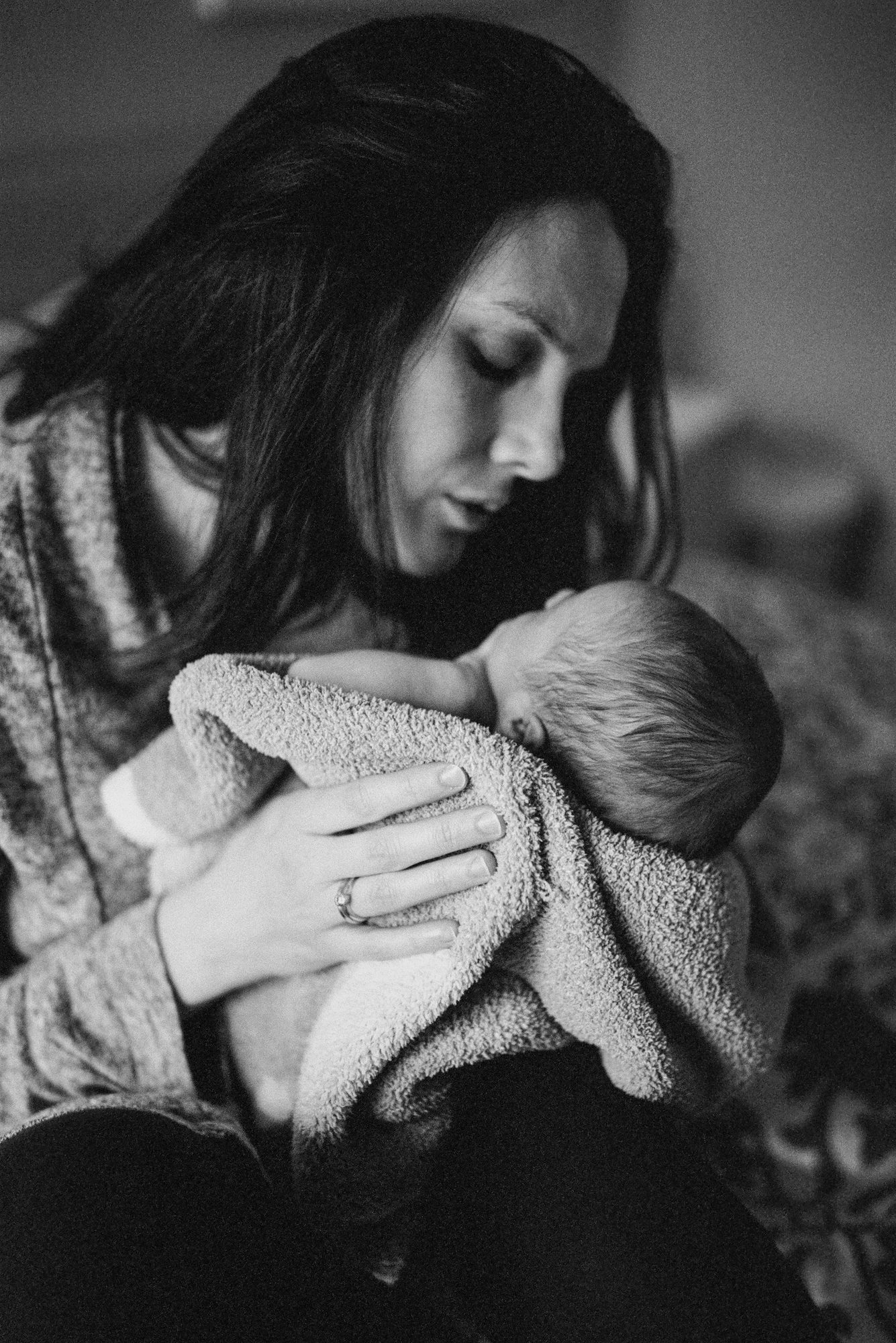 A mother tenderly cradles her newborn's head, wedding ring visible on her hand, in soft moody light