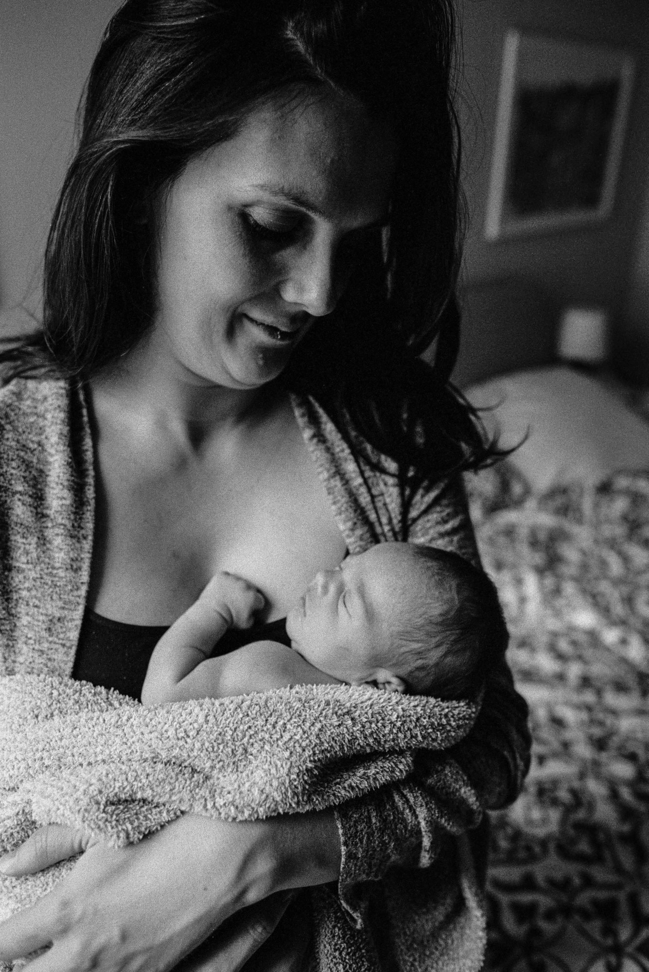 A mother smiles softly down at her nursing newborn, seated on the bed in warm natural light