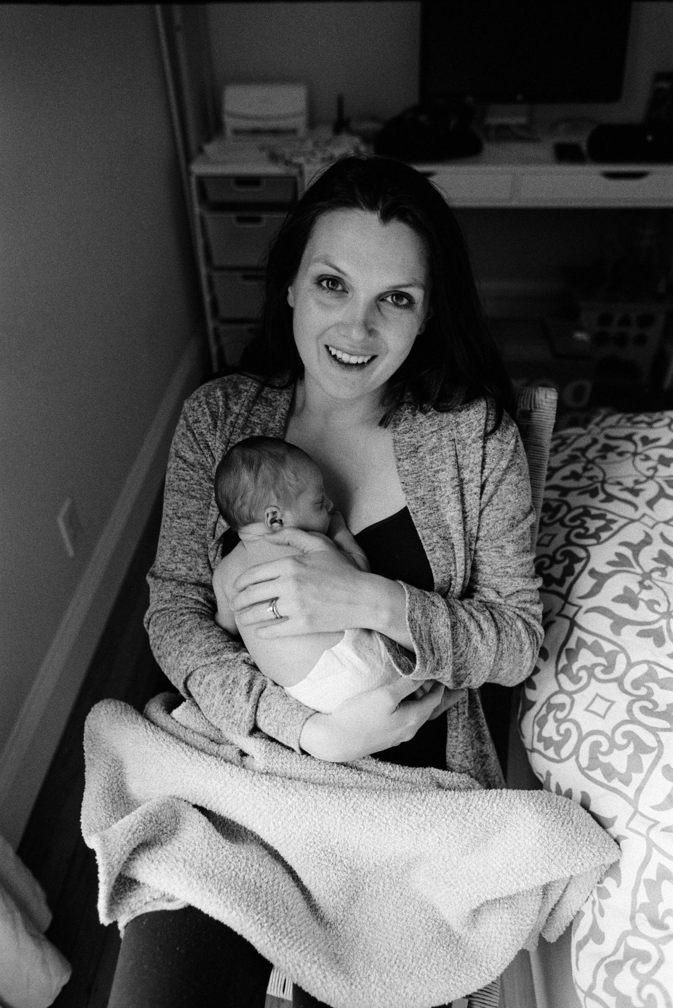 A joyful mother smiles at the camera while holding her swaddled newborn, seated beside the bed