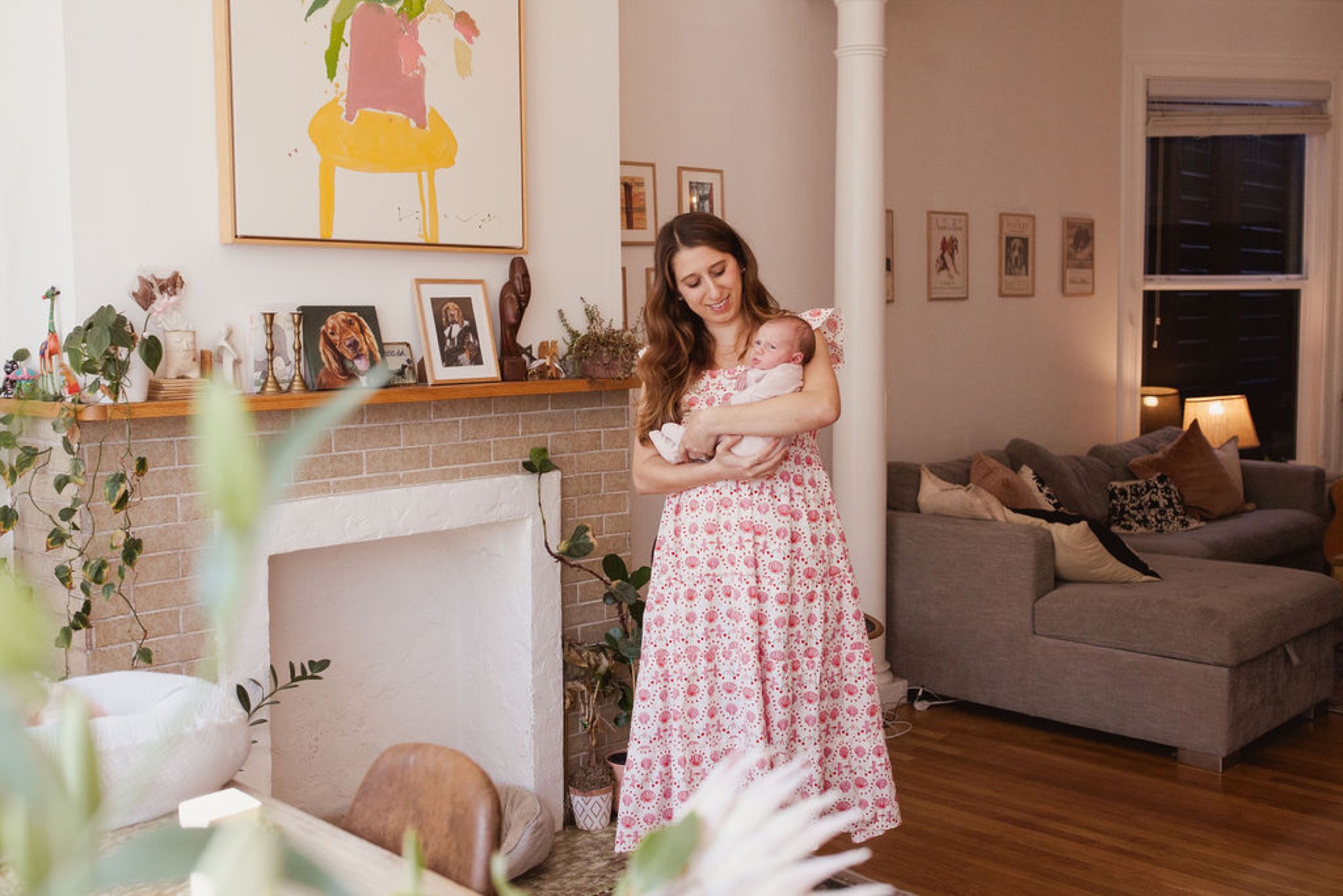 Mother in a pink floral dress holds her newborn against her chest while standing beside a brick fireplace adorned with family photos and greenery