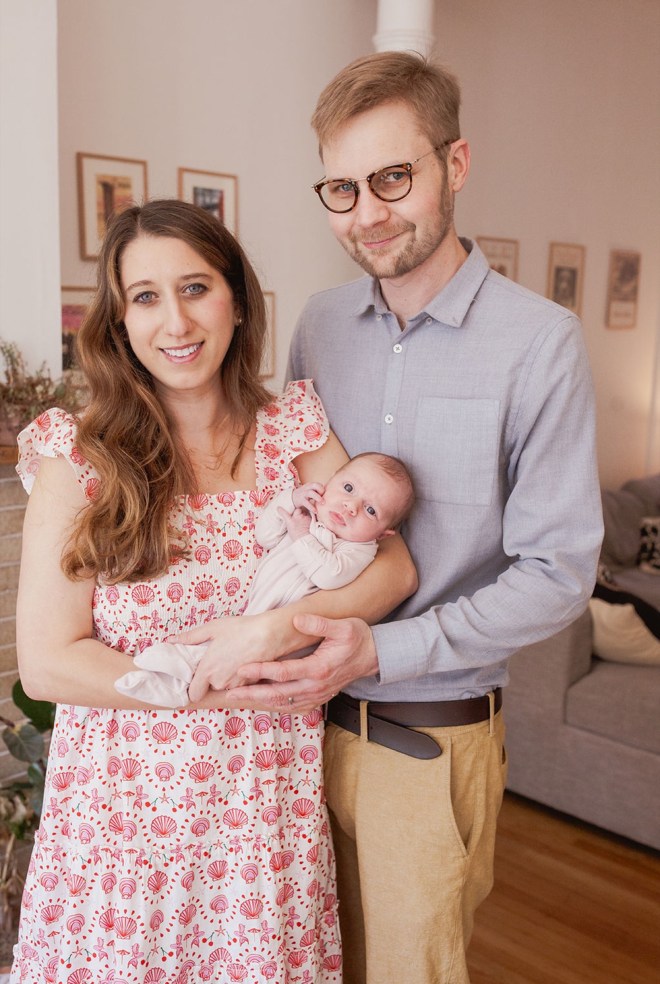 Parents beam at their newborn in a warm living room, the father's glasses reflecting soft natural light while holding their swaddled infant